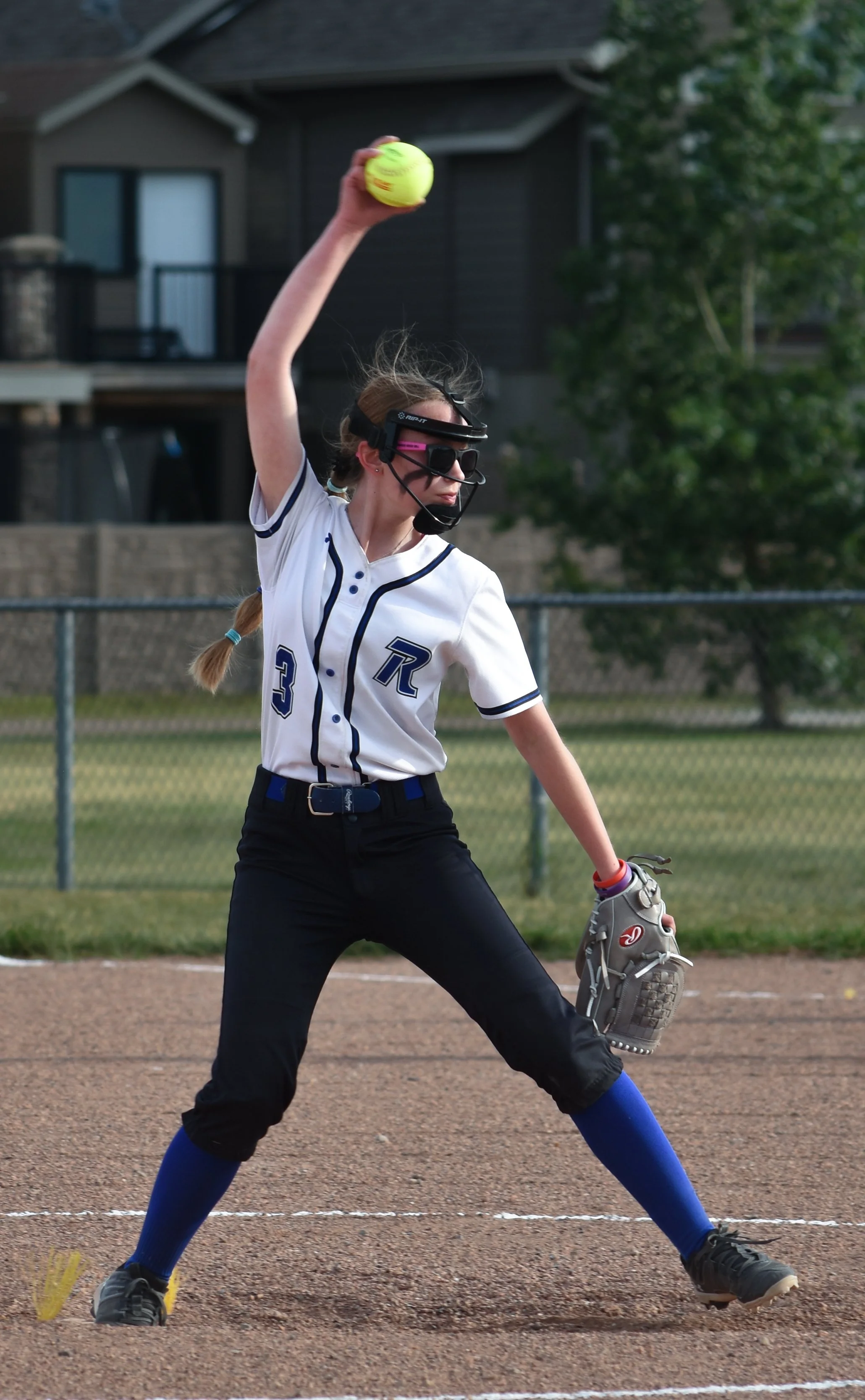 A female softball pitcher in uniform throwing a yellow softball on a field.