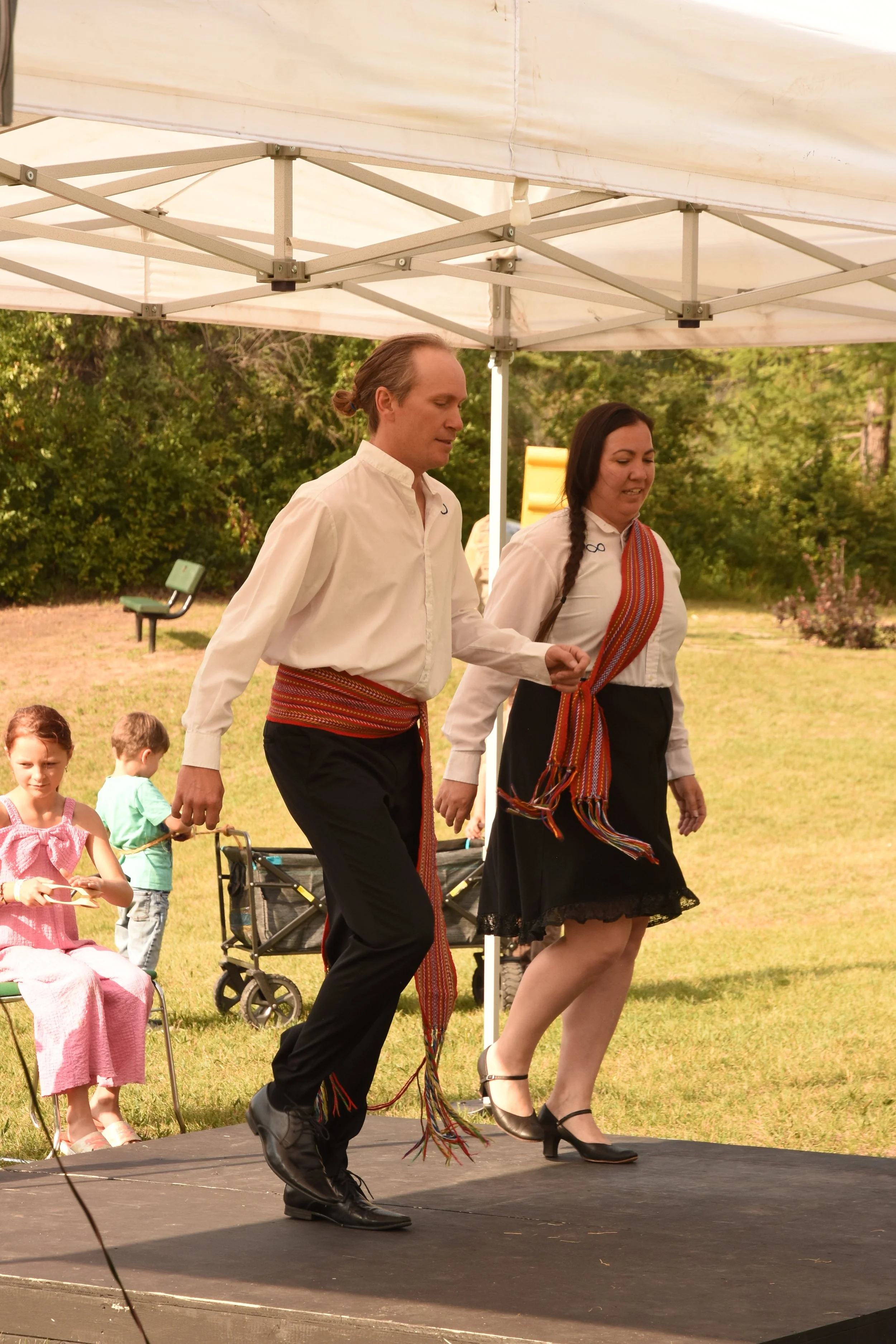 Man and woman in traditional clothing dancing on an outdoor stage under a canopy with children watching nearby.