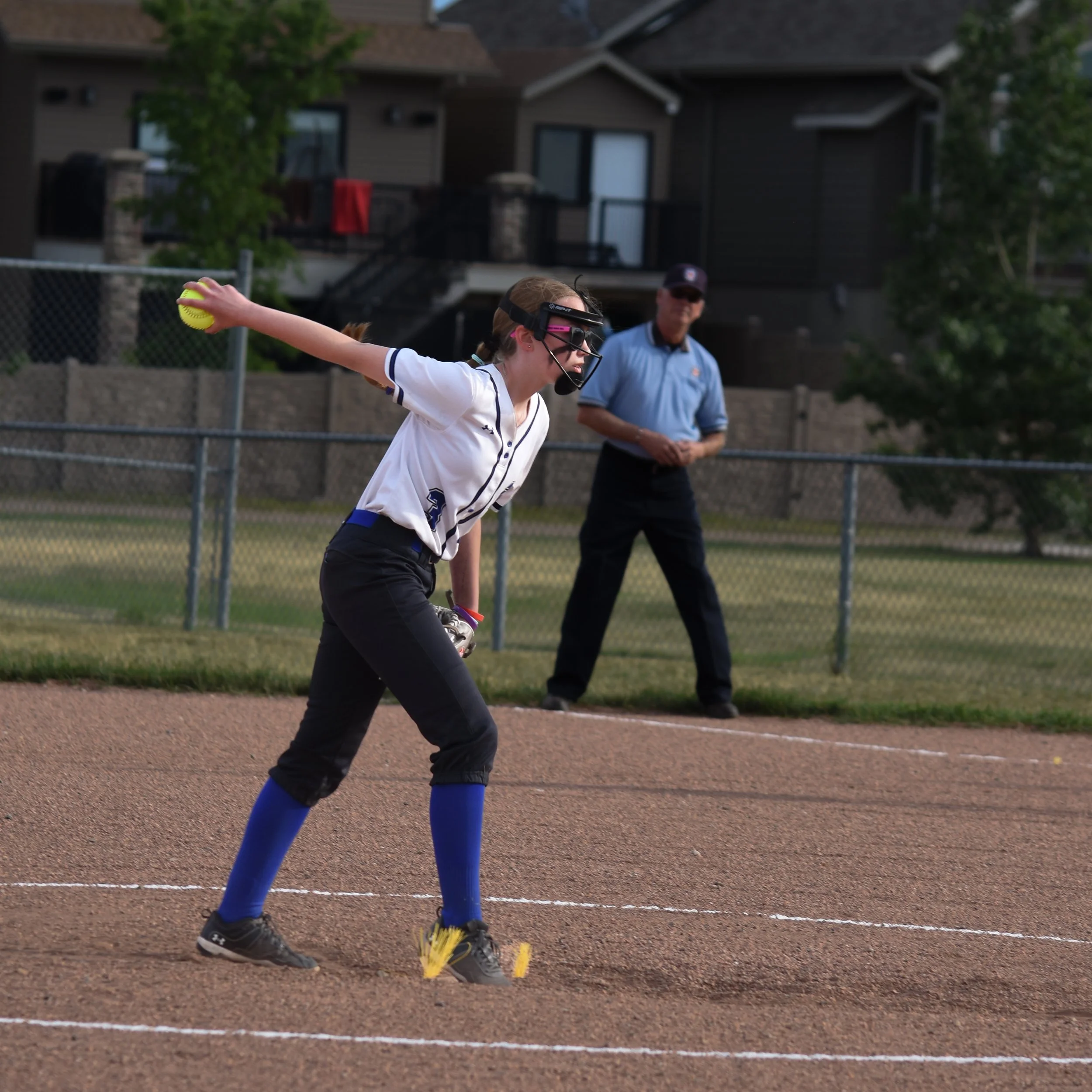 A female softball player in black pants, a white jersey, and blue socks is throwing a yellow softball on a dirt field, with an umpire standing in the background.