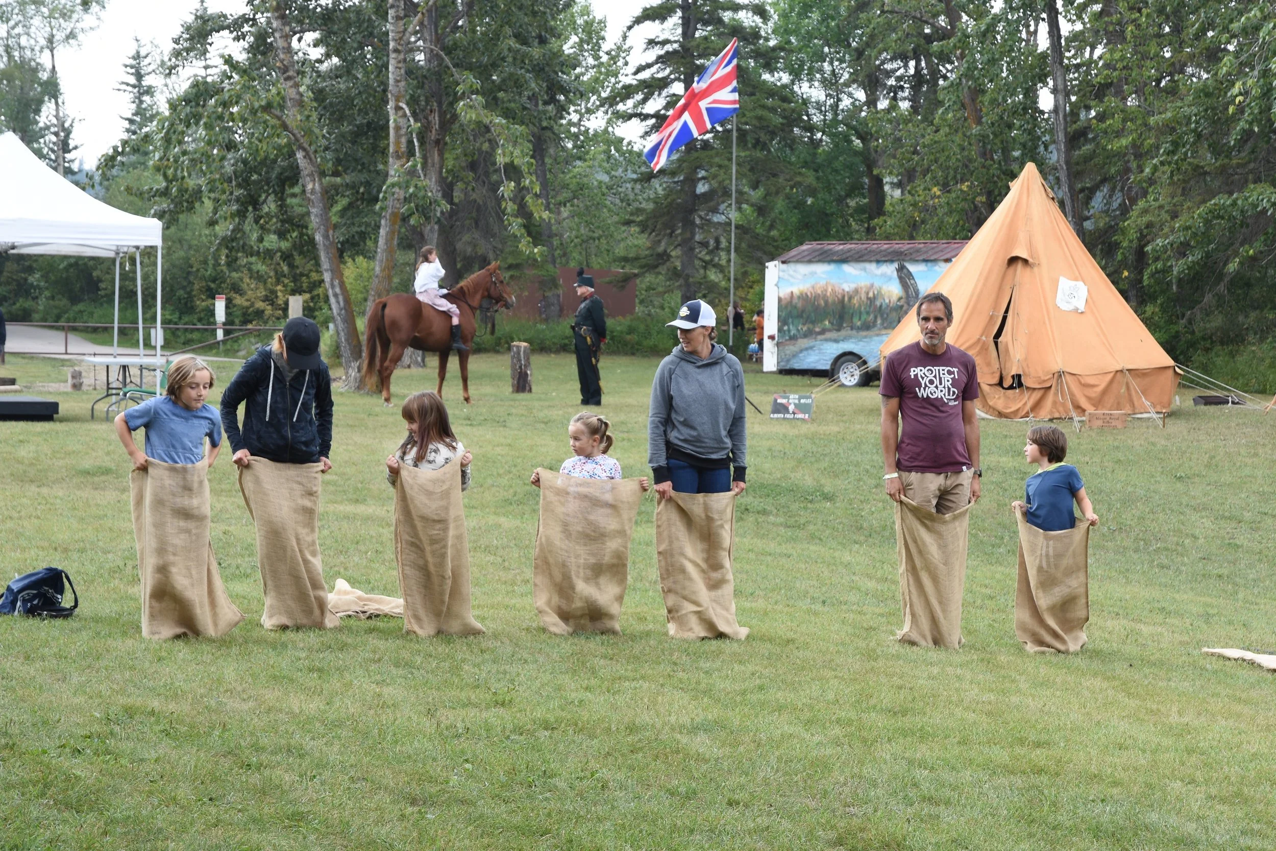 Children and adults participating in a sack race outdoors on a grassy field, with a teepee, Union Jack flag, trees, and a horse with a rider in the background.