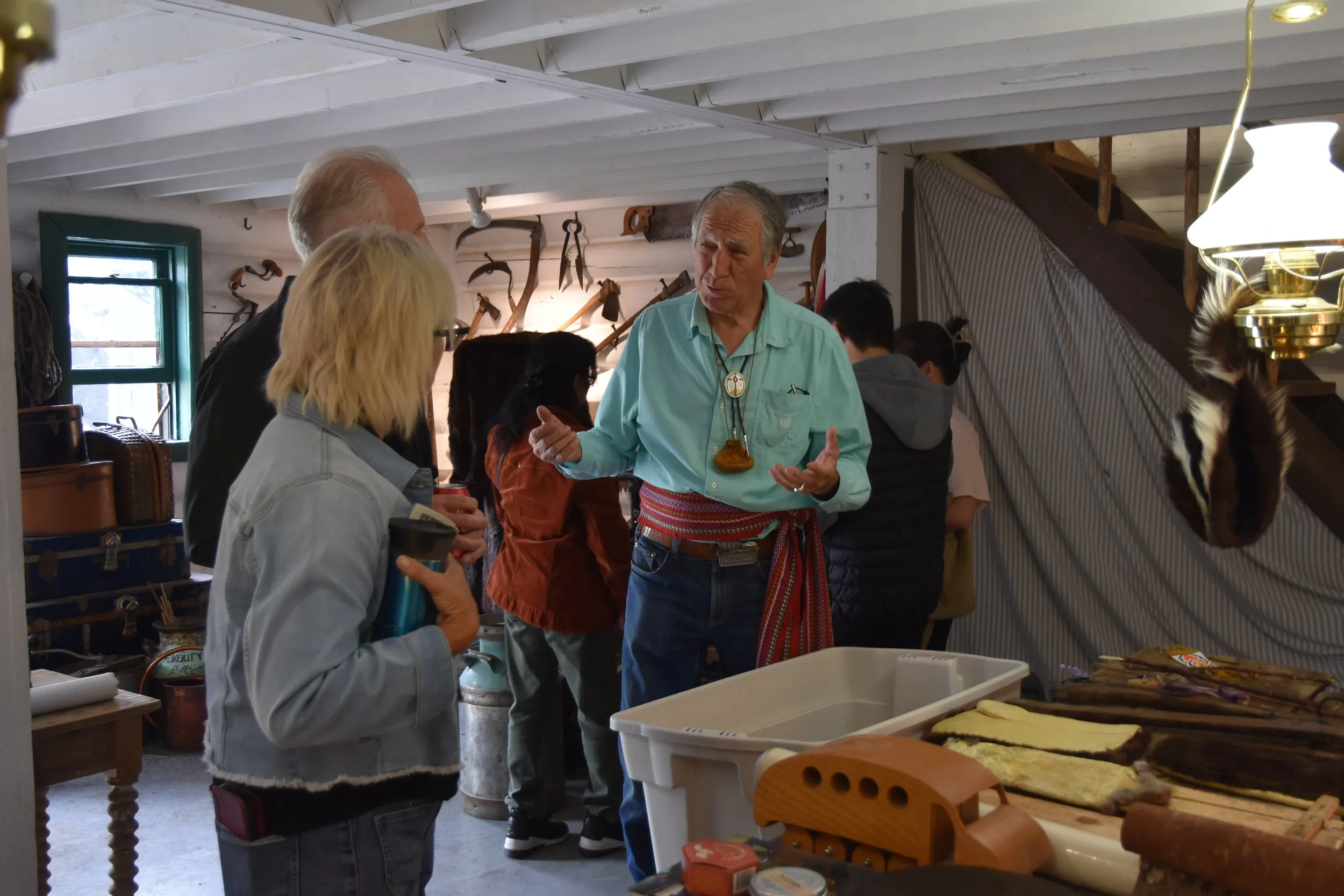A group of people gathered inside a rustic room, engaging in conversation with a man wearing a turquoise shirt and a bolo tie. The room has wooden beams, tools hanging on the wall, and several tables with food and supplies.