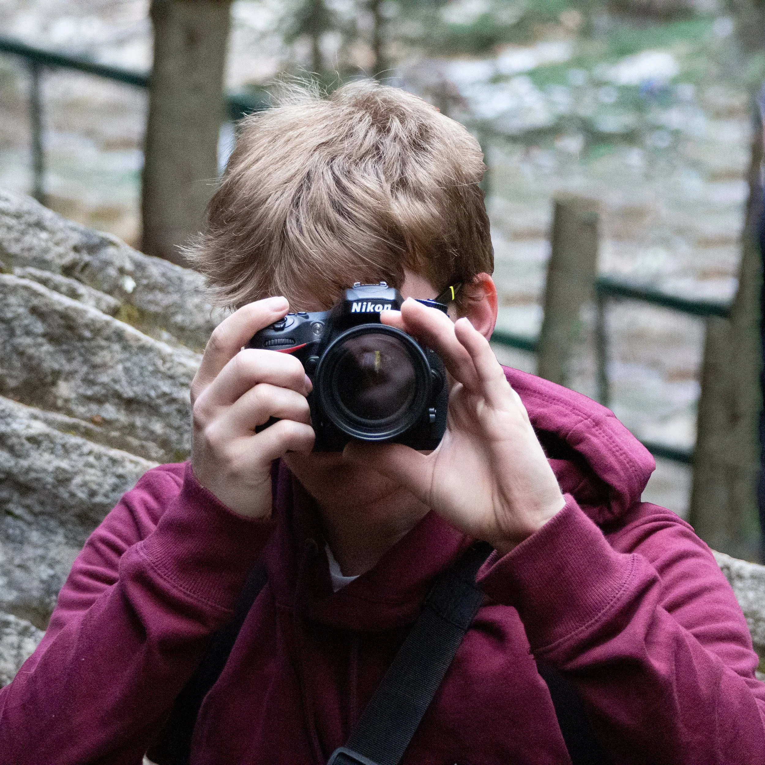 A person with brown hair wearing a maroon hoodie and glasses takes a photo with a Nikon DSLR camera in an outdoor setting with trees and rocks.