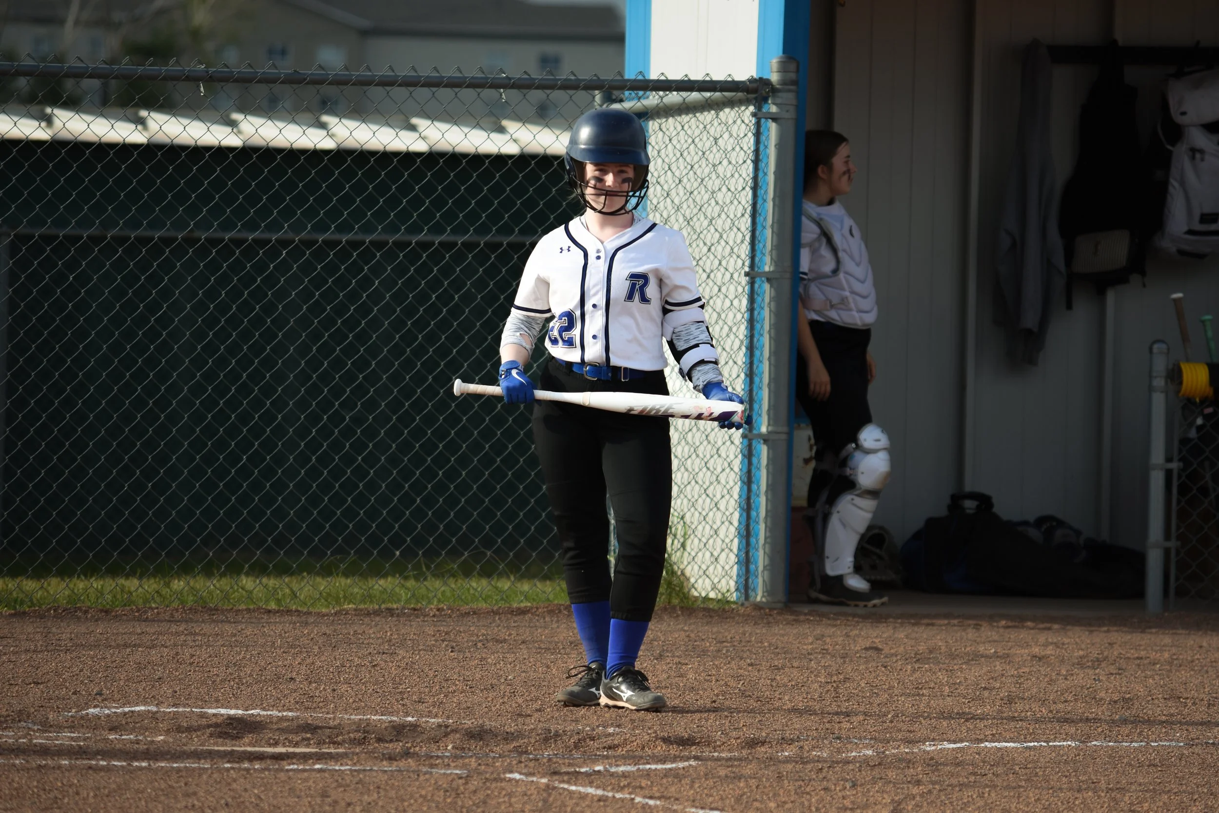A young female softball player standing on the dirt field holding a bat, wearing a helmet, jersey, and gloves, with another player in catcher's gear in the dugout in the background.