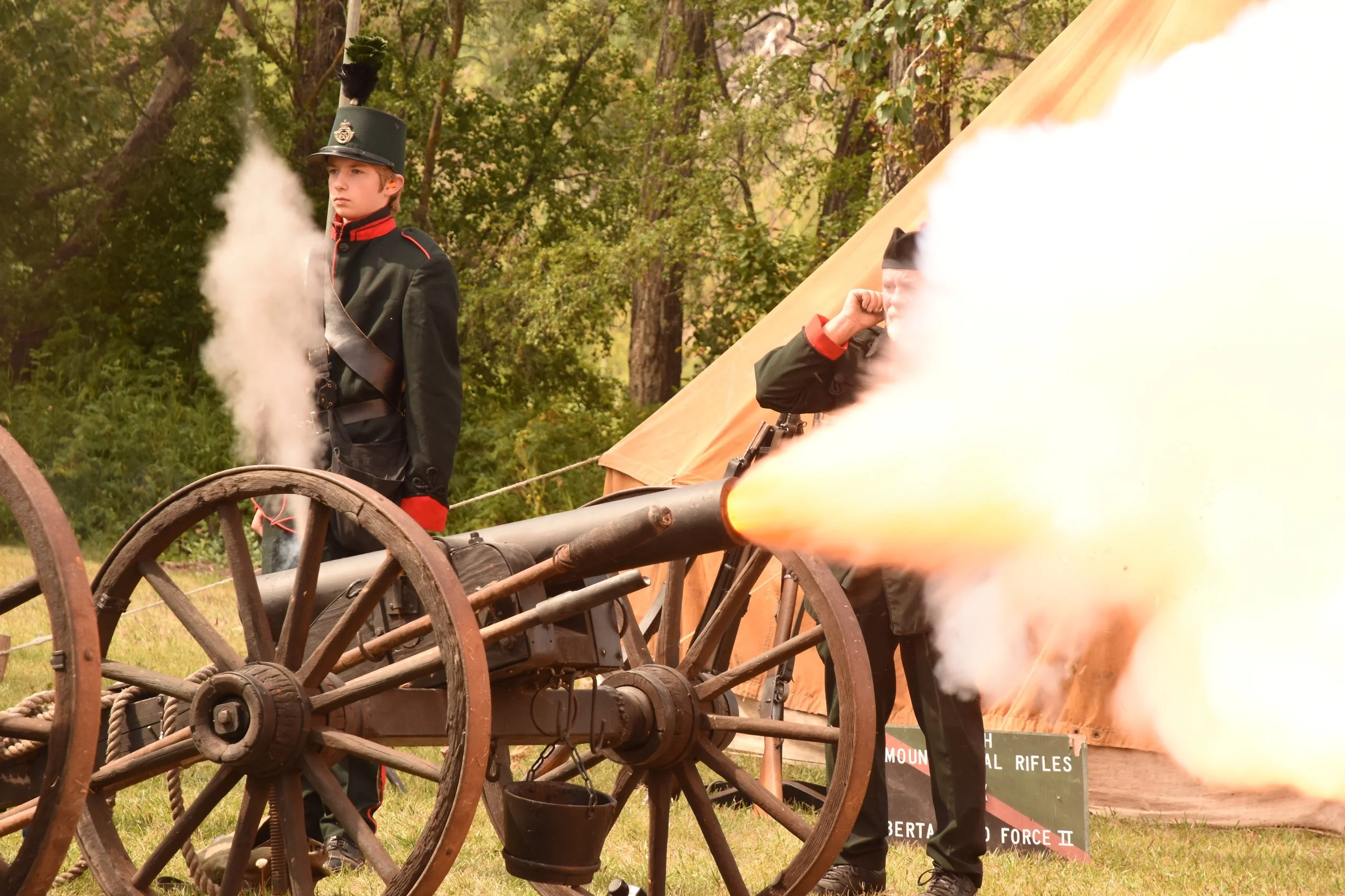 Two young men dressed in historical military uniforms operating an old-fashioned cannon that is firing, with smoke coming out of the barrel. They are outdoors, with green trees and a tent in the background.