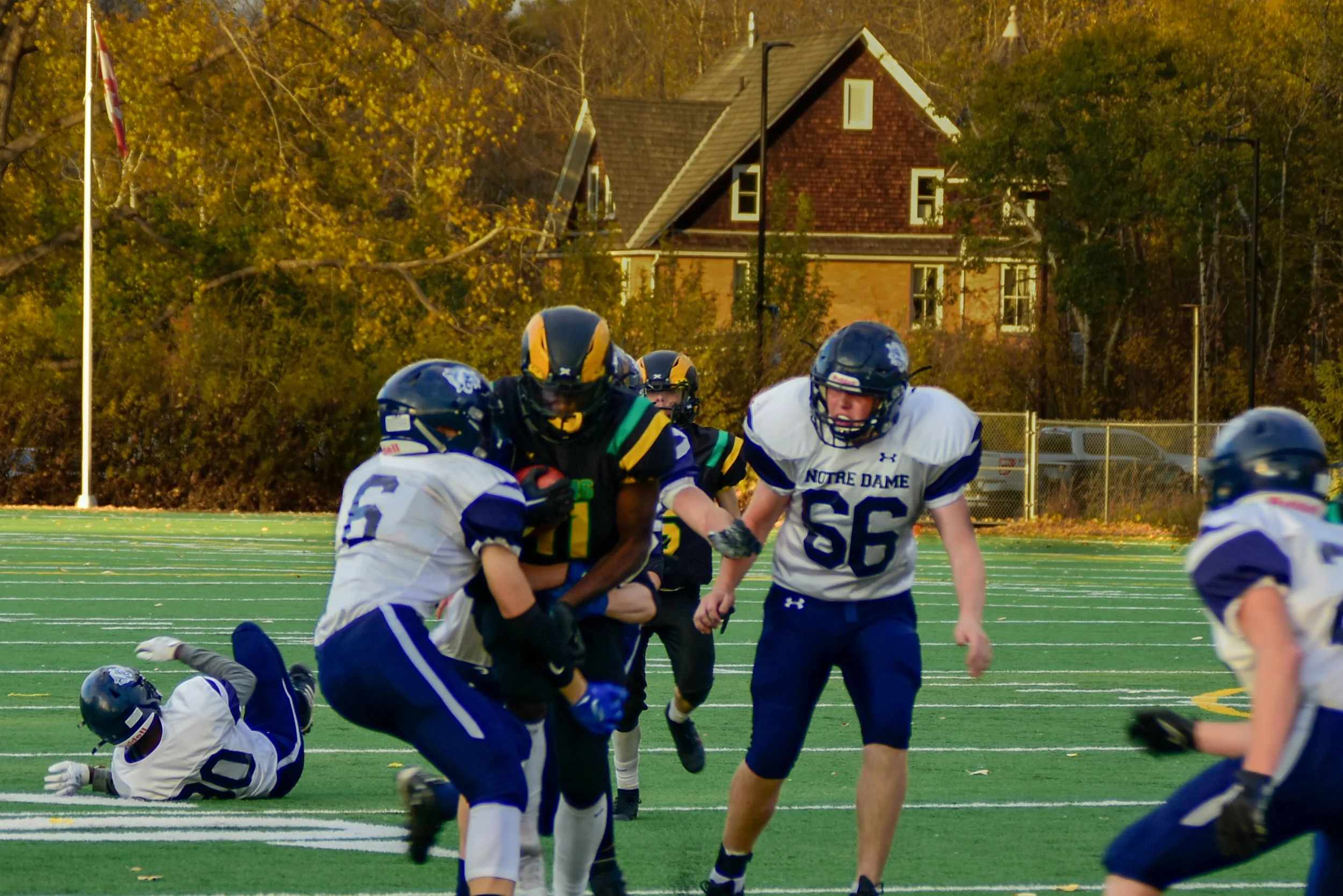 American football game with players from Notre Dame and another team on a green field with autumn trees and houses in the background.