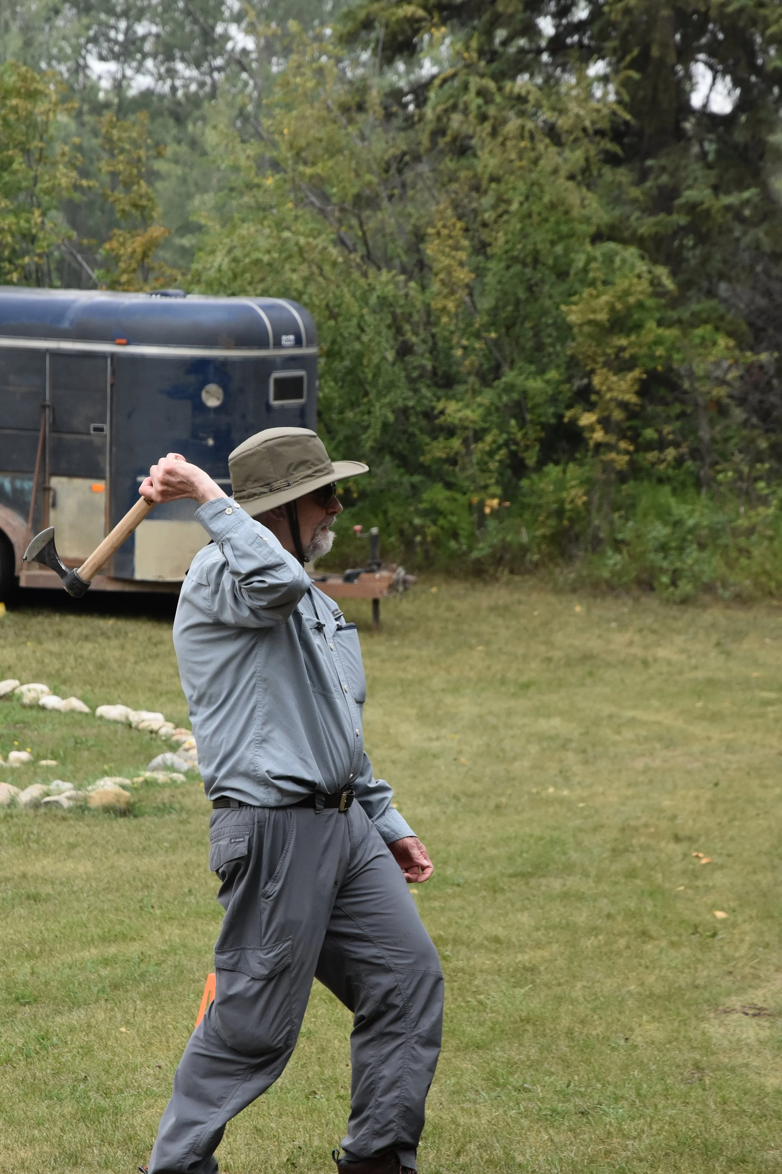 An elderly man in outdoor gear, including a wide-brimmed hat, sunglasses, and gray clothing, holds an axe over his shoulder while standing on a grassy area near trees and a blue trailer, possibly in a park or backyard.
