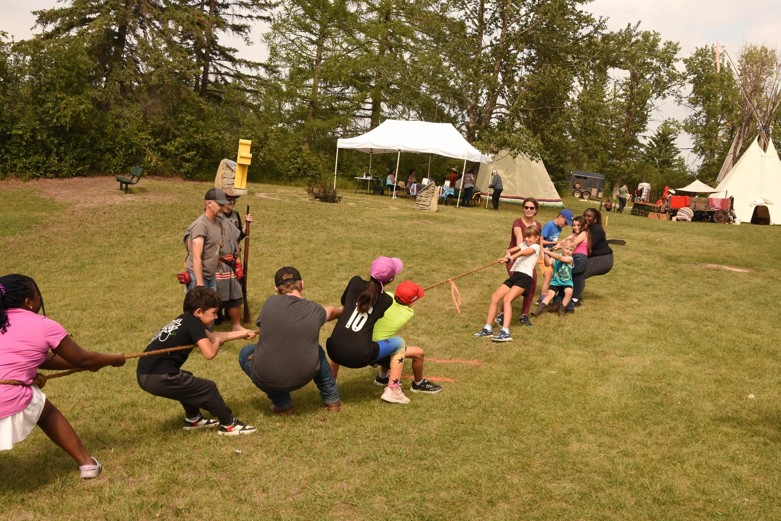 Children and adults participate in a tug-of-war game on a grassy field with tents and trees in the background, at an outdoor event.