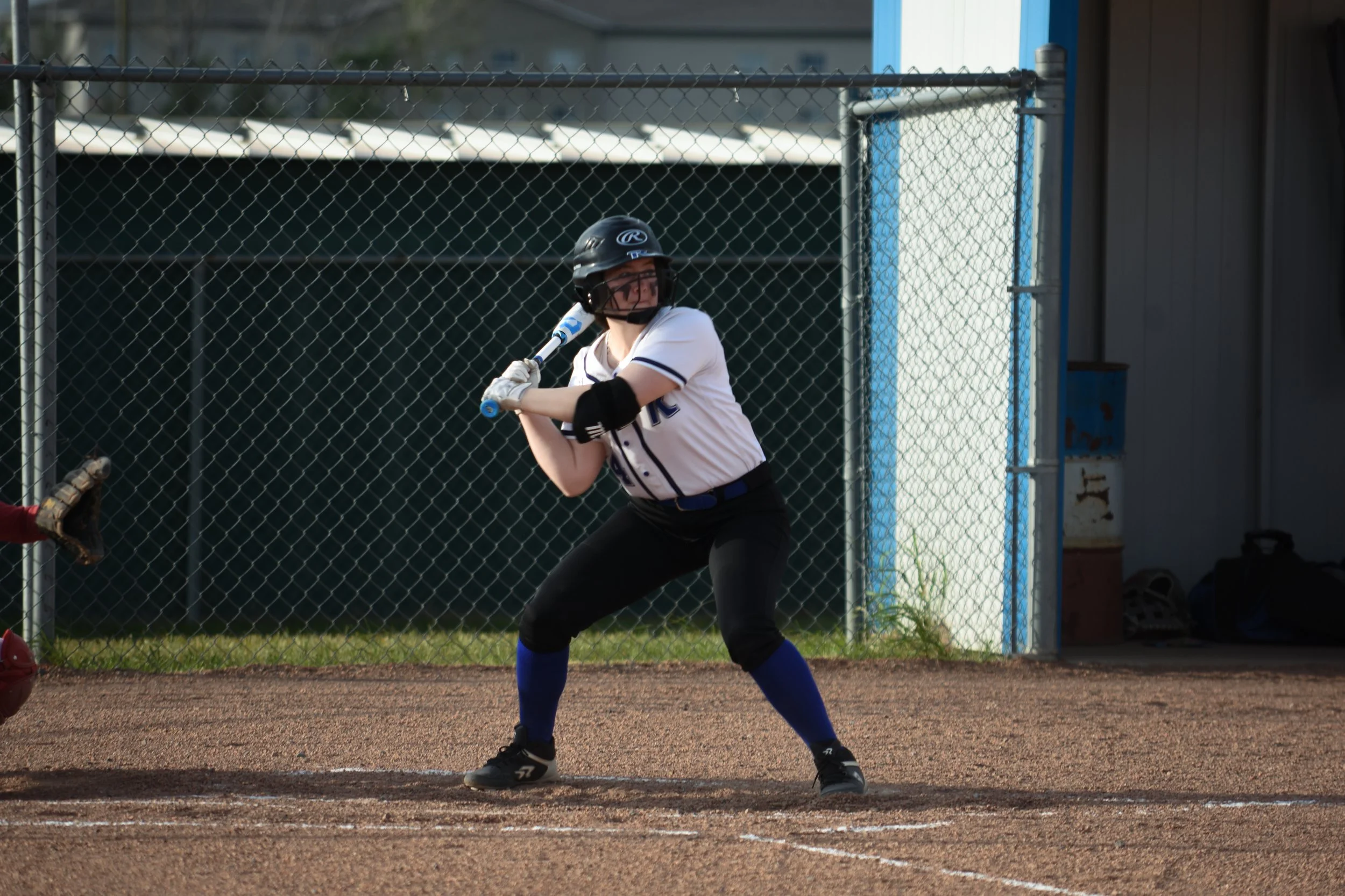 Girl in baseball uniform stands at home plate with bat ready to swing on softball field.