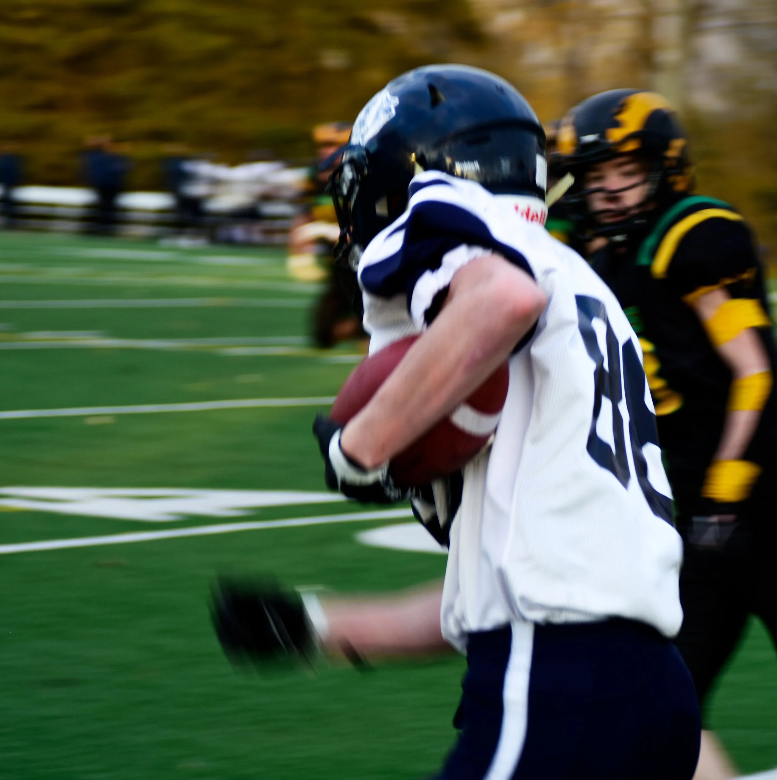 Football player running with the ball during a game, wearing a white jersey and black helmet, with an opponent in black and yellow on the field.