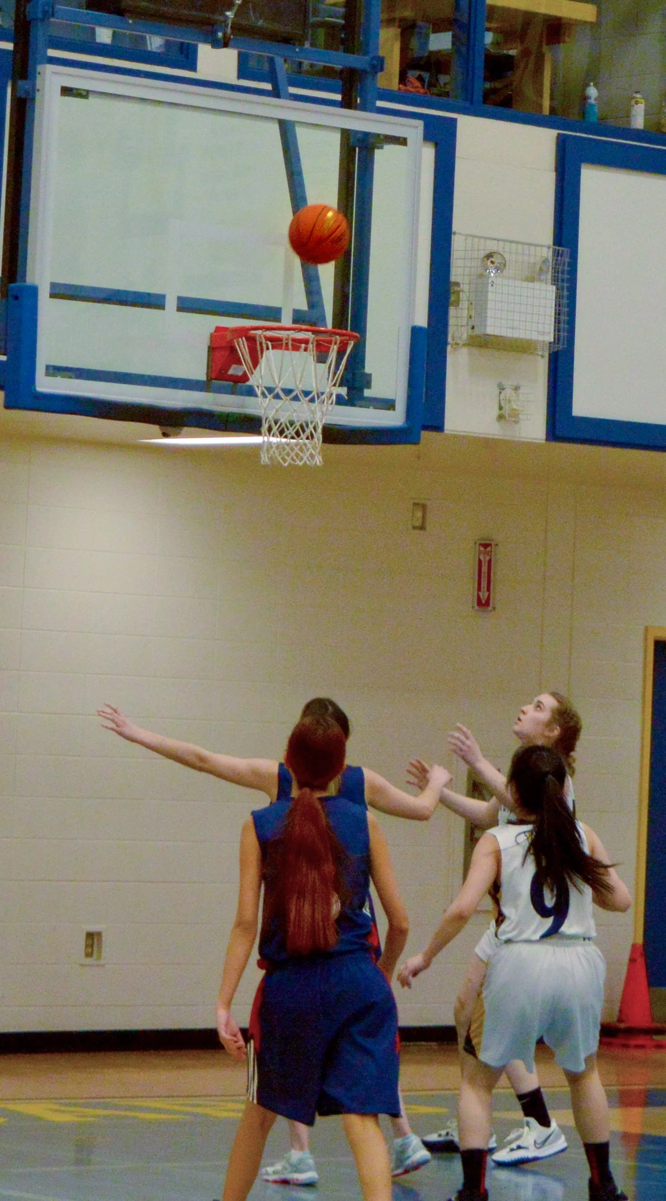 A basketball game in progress with four players near the basket, one shooting the ball towards the hoop, with the basketball mid-air near the basket, inside a gymnasium.