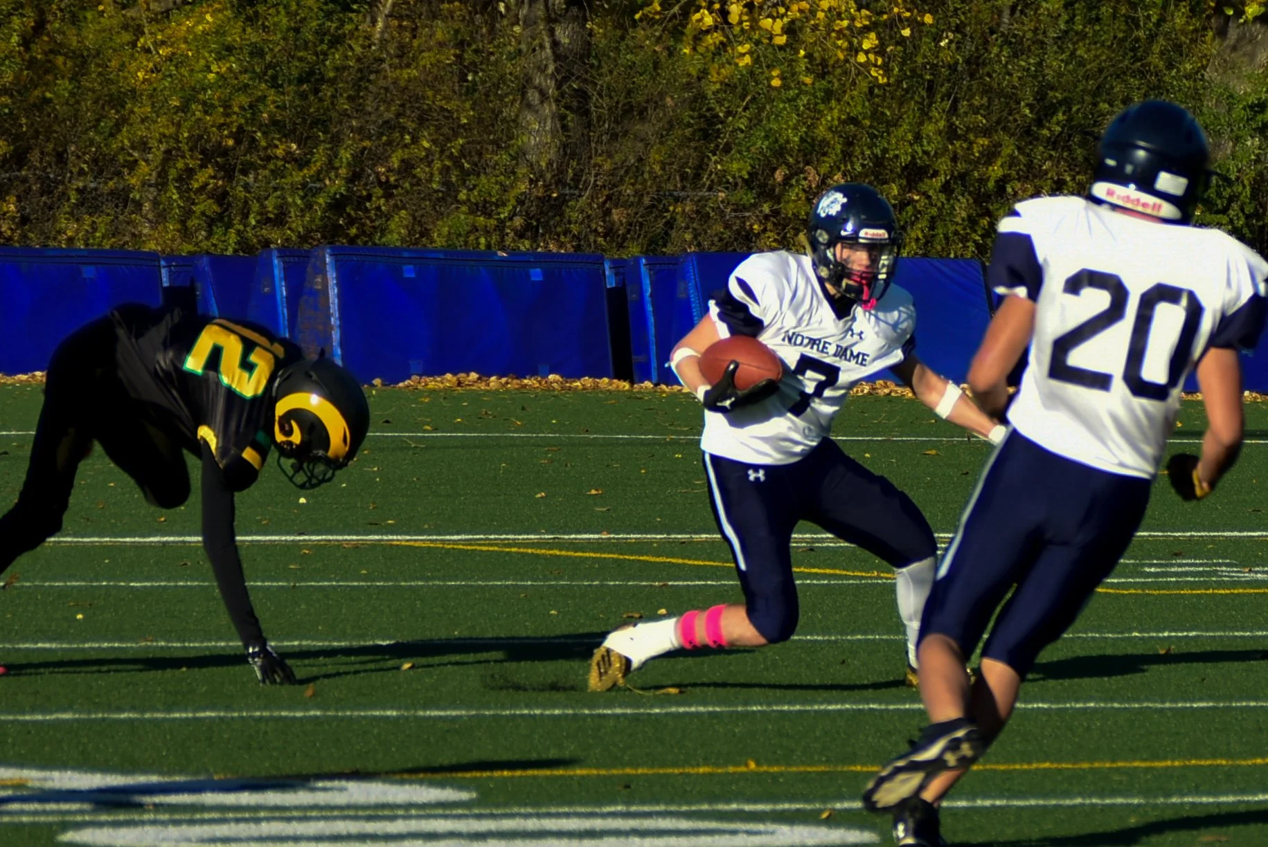 American football players during a game, with one player running with the ball and two others attempting to block or tackle, on a green field with white yard lines and a blue boundary fence in the background.
