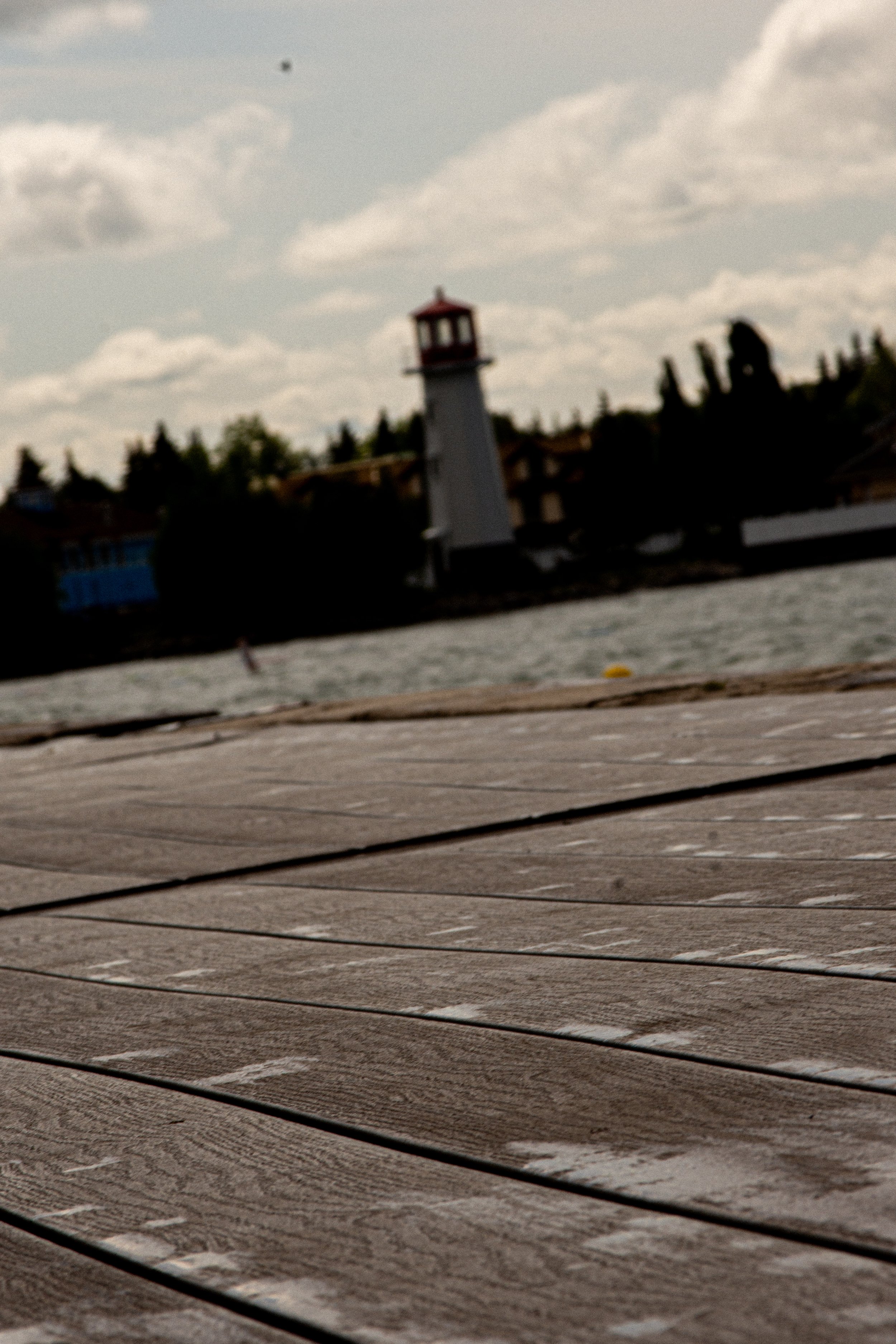 Close-up of a wooden dock or boardwalk near a body of water, with a distant lighthouse and houses in the background under a cloudy sky.