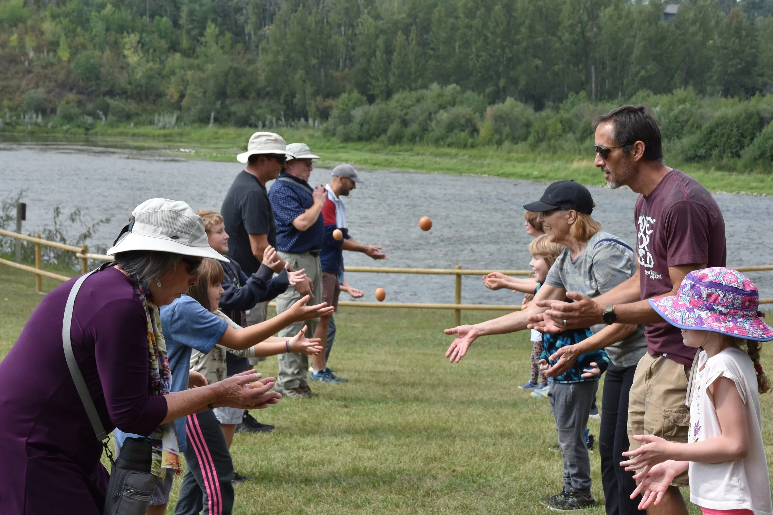 People outdoors near a lake playing a game involving tossing or catching balls, with children and adults participating.
