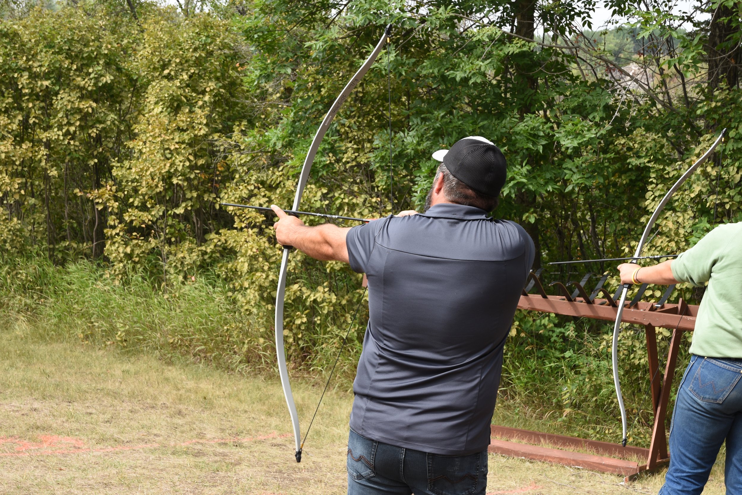 Two people practicing archery outdoors, one man aiming with a bow and arrow, and another person holding a bow. They are in a grassy area with trees in the background.