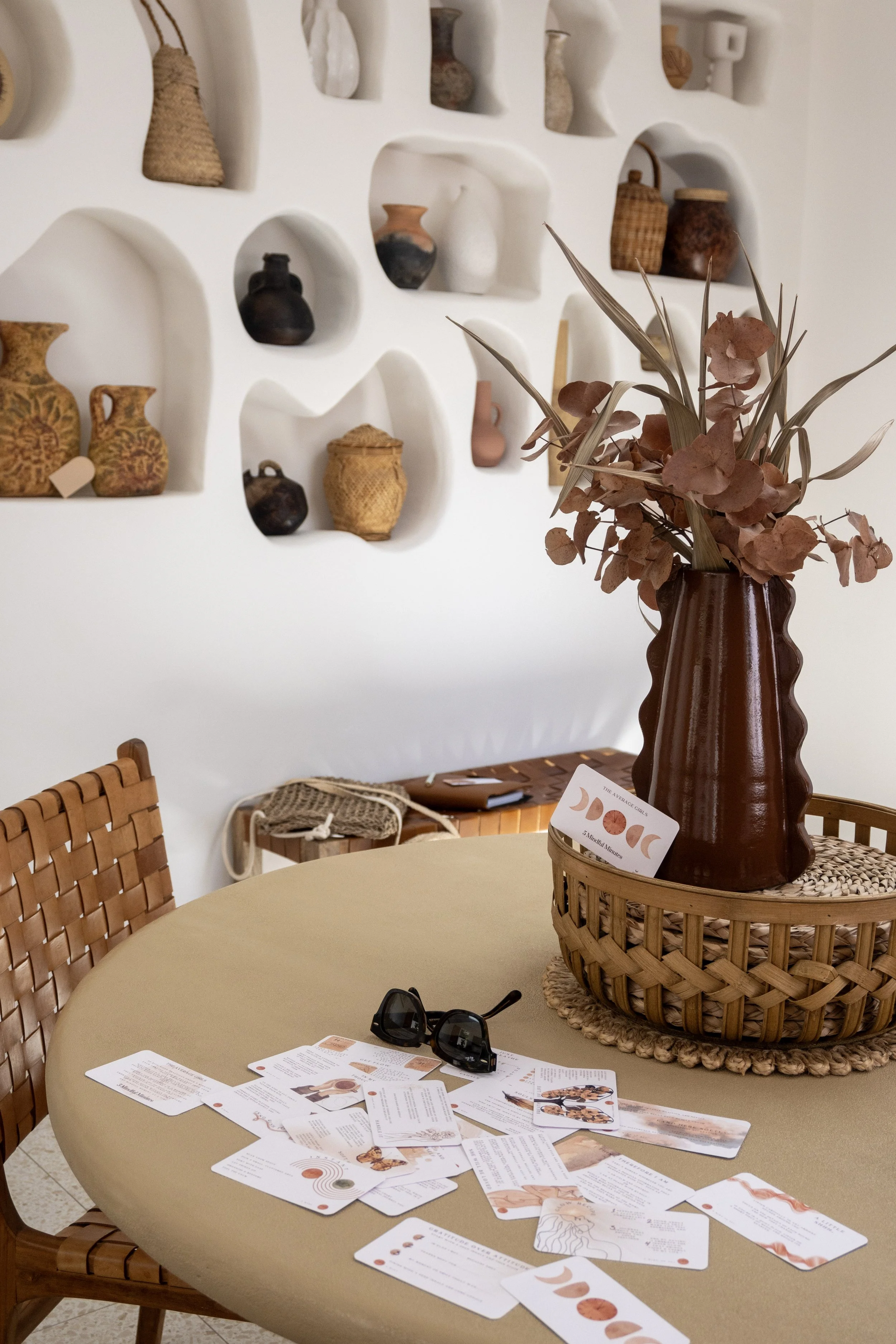 Interior space with a round table, scattered cards, sunglasses, vase with dried flowers, woven chair, and decorative wall alcoves with pottery.