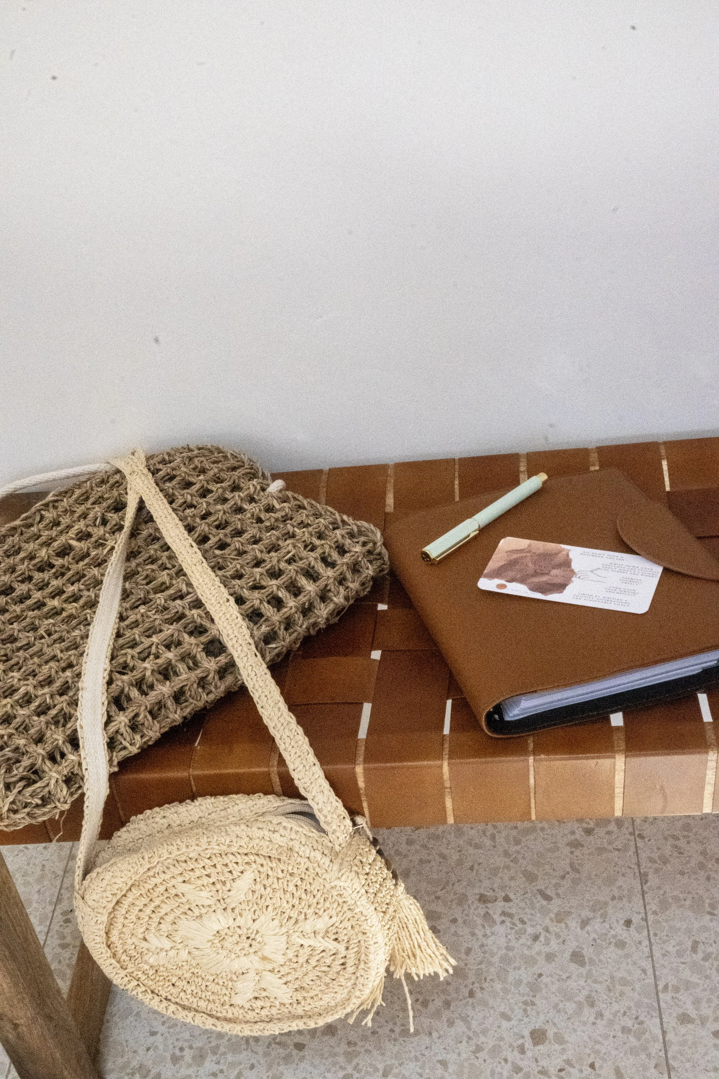 Woven bench with round straw bag, knitted tote, and brown folder holding pen and card.