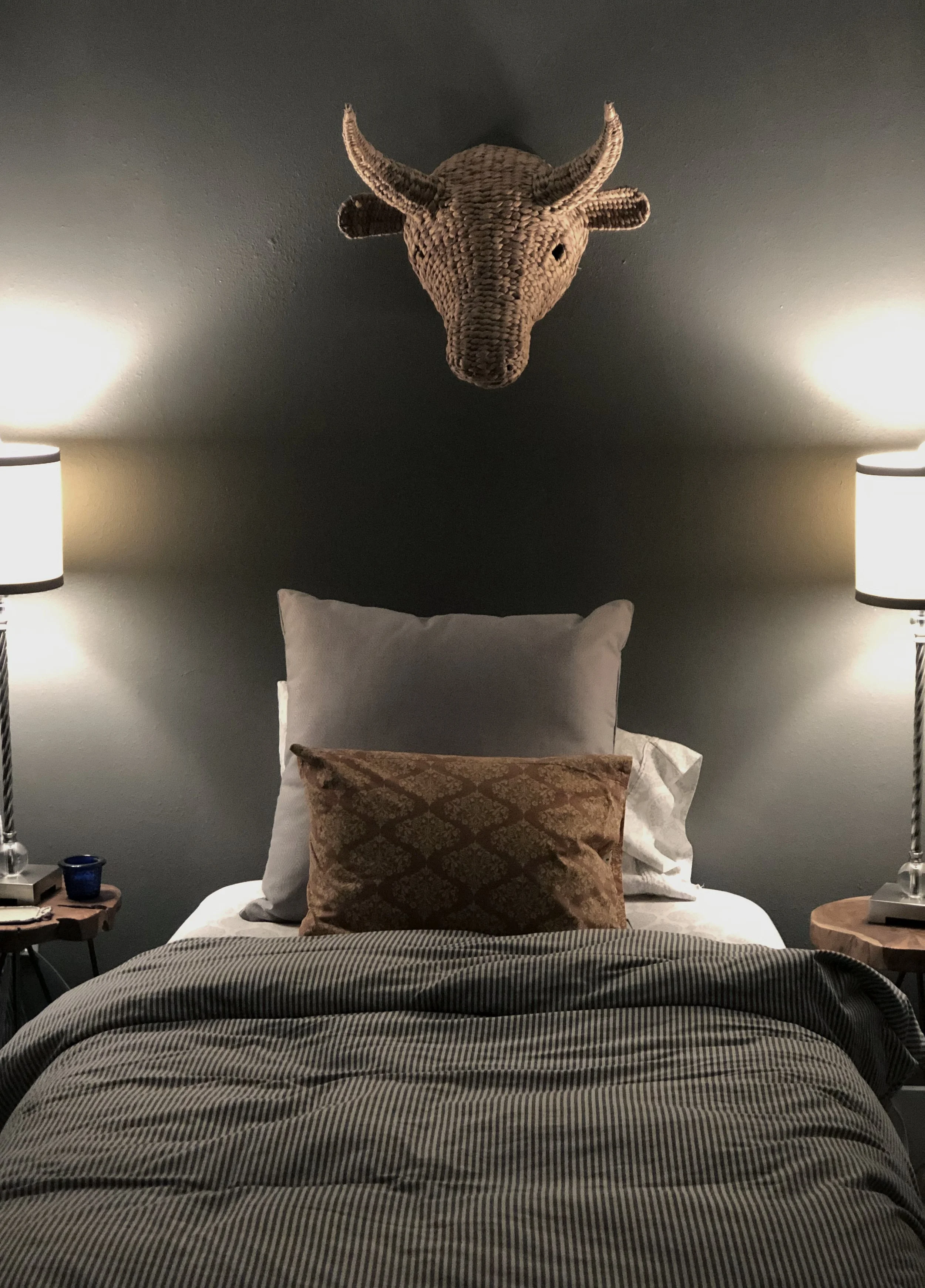 A small guest room with a twin bed is painted a deep warm grey in the same family as the grey throughout the rest of the home. Little pops of color appear in acacia nightstands, some complimentary pillows, a patterned area rug, and a basket-weave bul