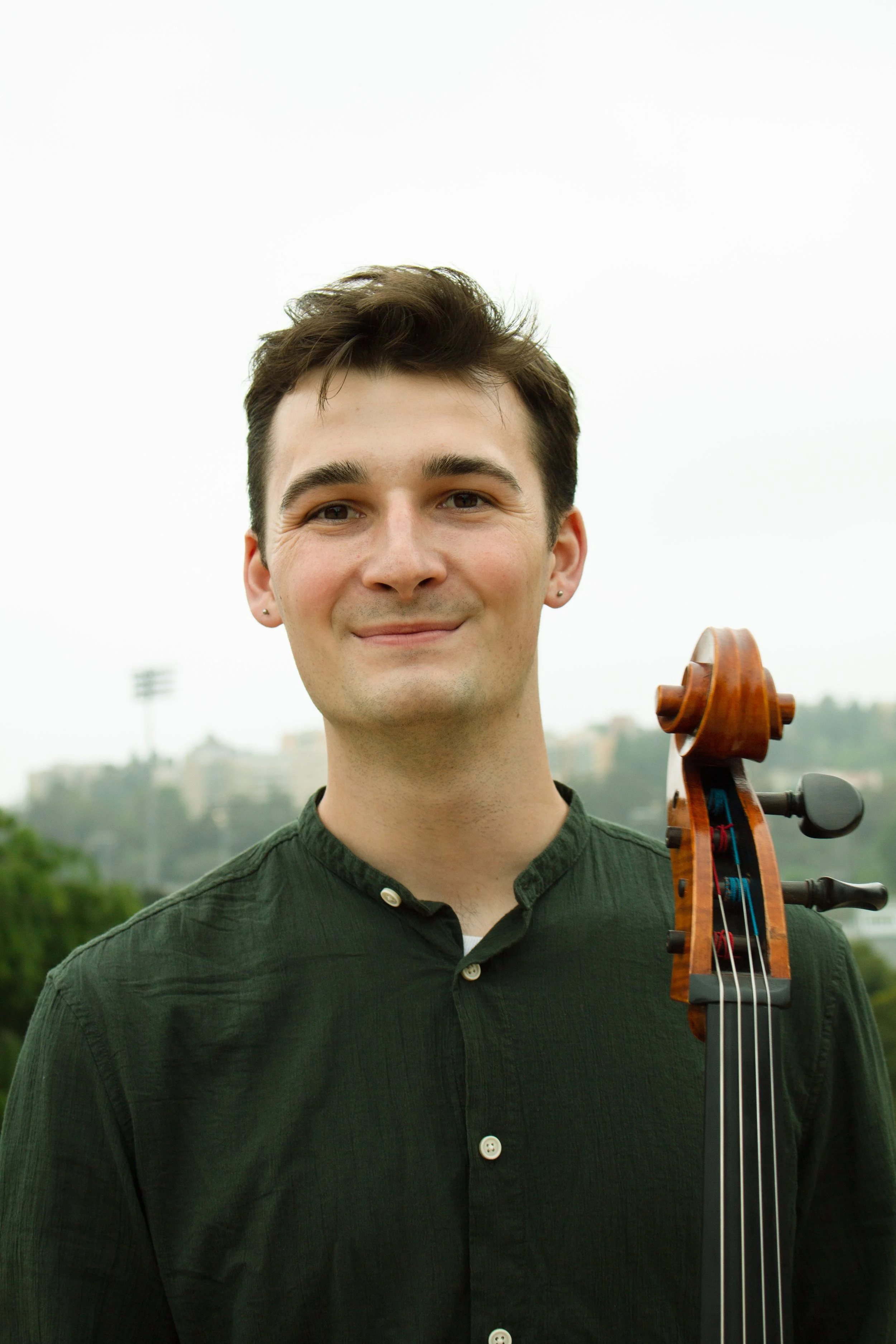Young man with brown hair smiling, wearing a dark green button-up shirt, holding a violin, outdoors in a park with trees and a cloudy sky. Nice Cello Teacher in Santa Clarita