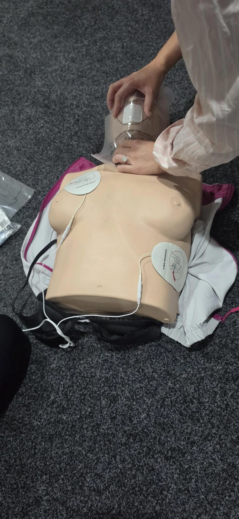 A medical training mannequin of a human torso lying on a gray carpeted floor, with a person performing CPR or medical procedure on it, wearing a white shirt with rolled-up sleeves.