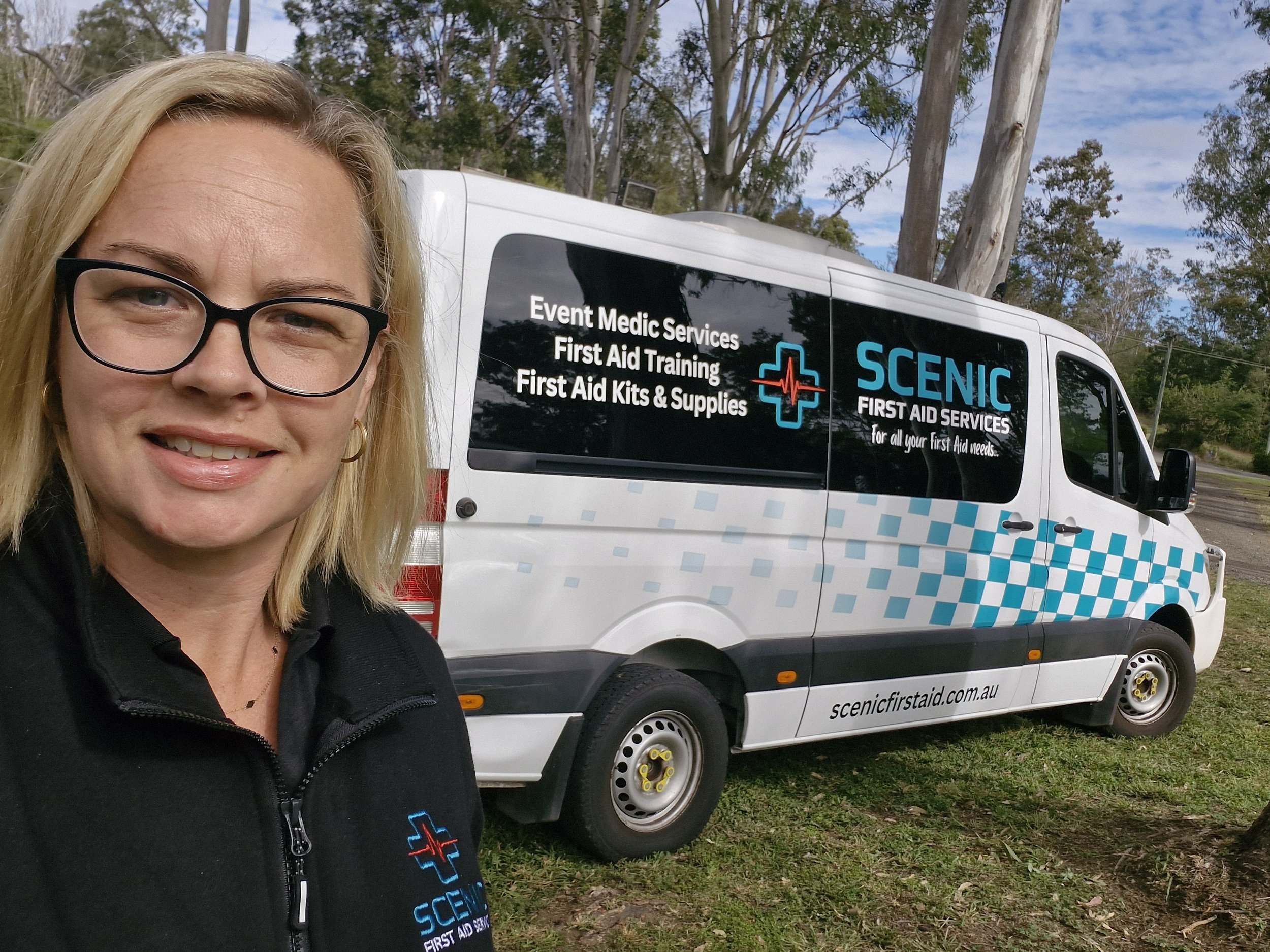 A woman with blonde hair and glasses smiling in front of a white vehicle with First Aid Service branding, including a red cross and medical symbols, parked outdoors with trees and blue sky in the background.