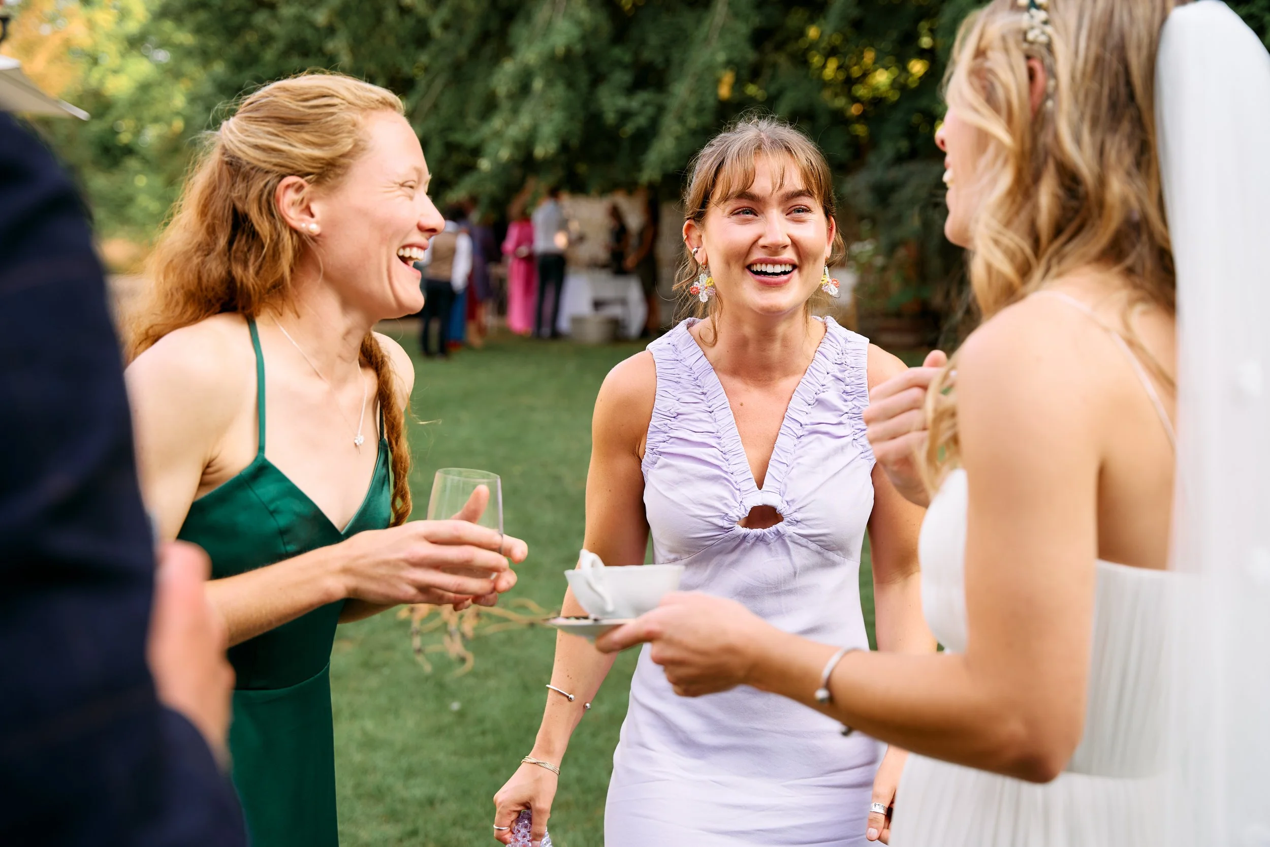 Three women laughing and talking at an outdoor event, with a woman in a white dress holding a tray offering drinks.