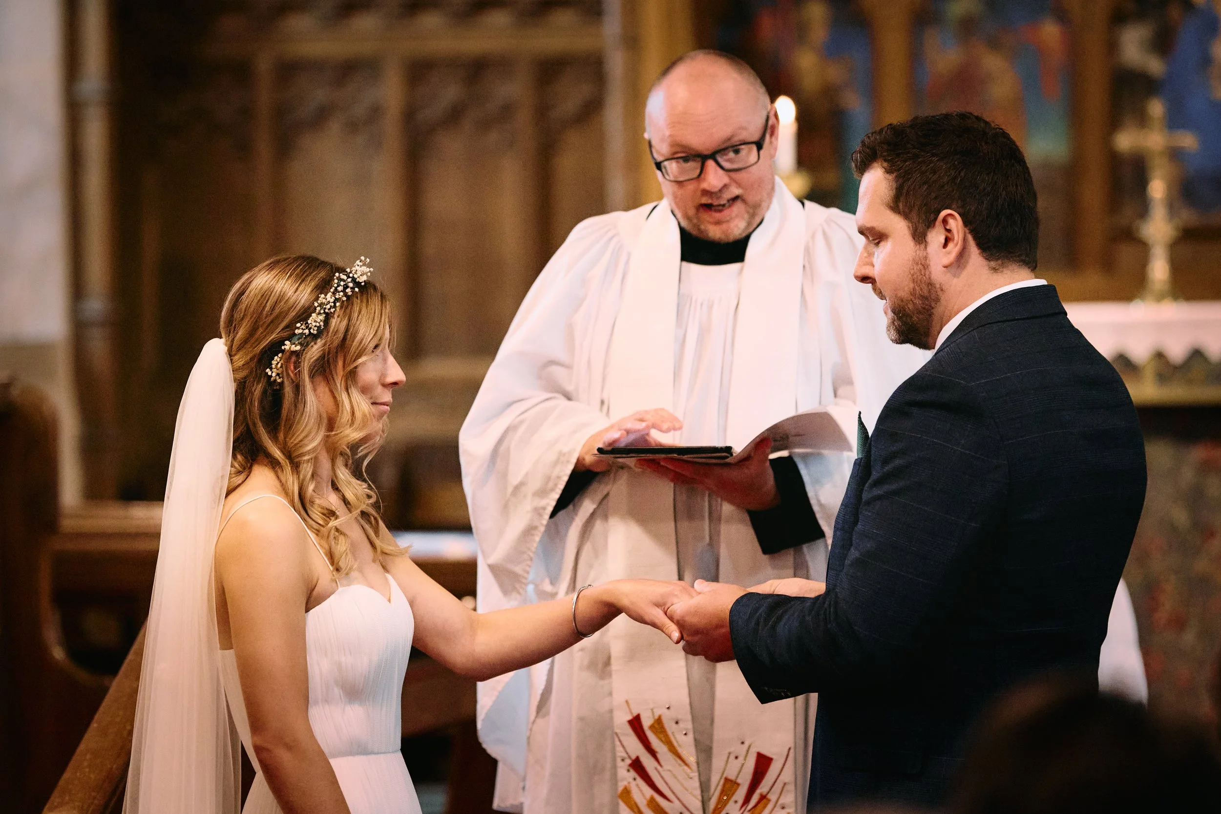 A bride and groom holding hands during their wedding ceremony in a church, with a priest officiating and holding a book.