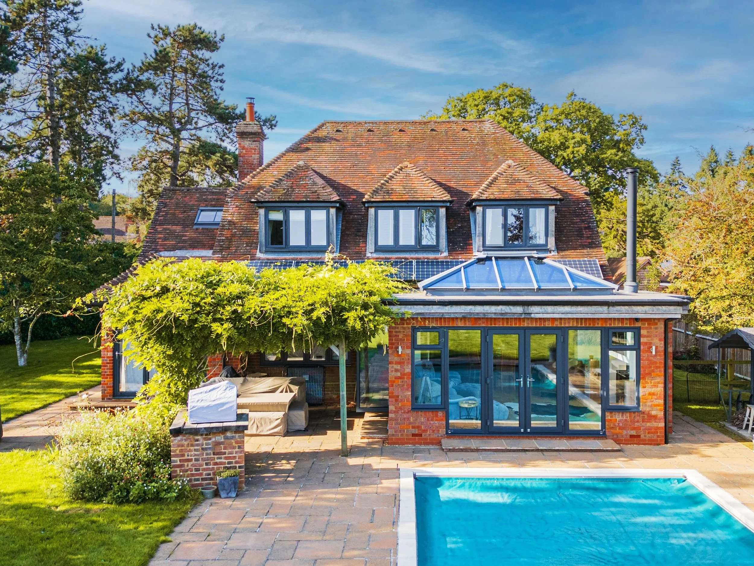 Rear view of a brick house with a swimming pool in the backyard, outdoor seating area, lush green trees, and a clear blue sky.