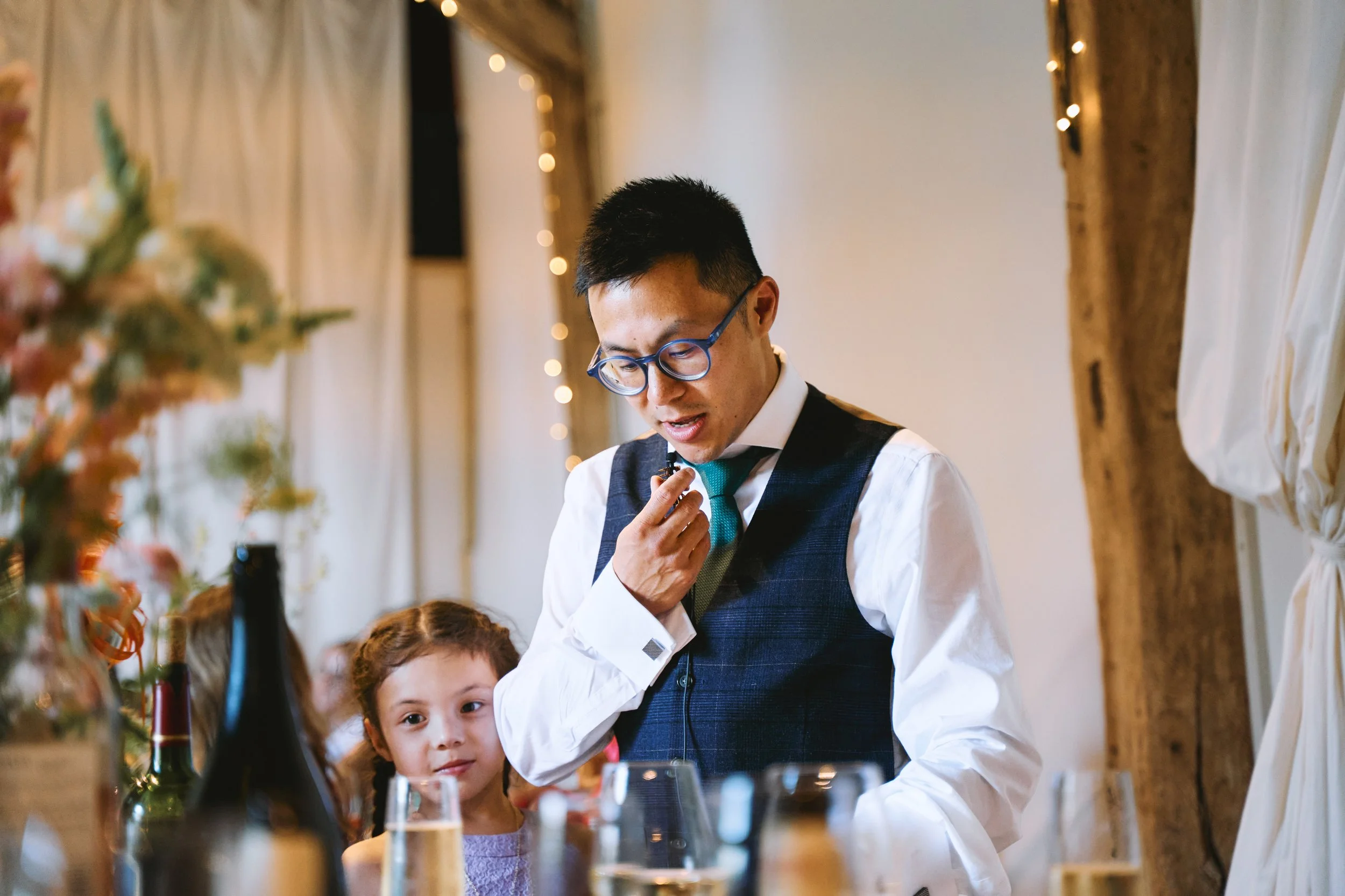A man in glasses and a vest, looking at something with a thoughtful expression, and a young girl watching him, during a gathering with bottles and glasses on the table, decorated with fairy lights and curtains in the background.
