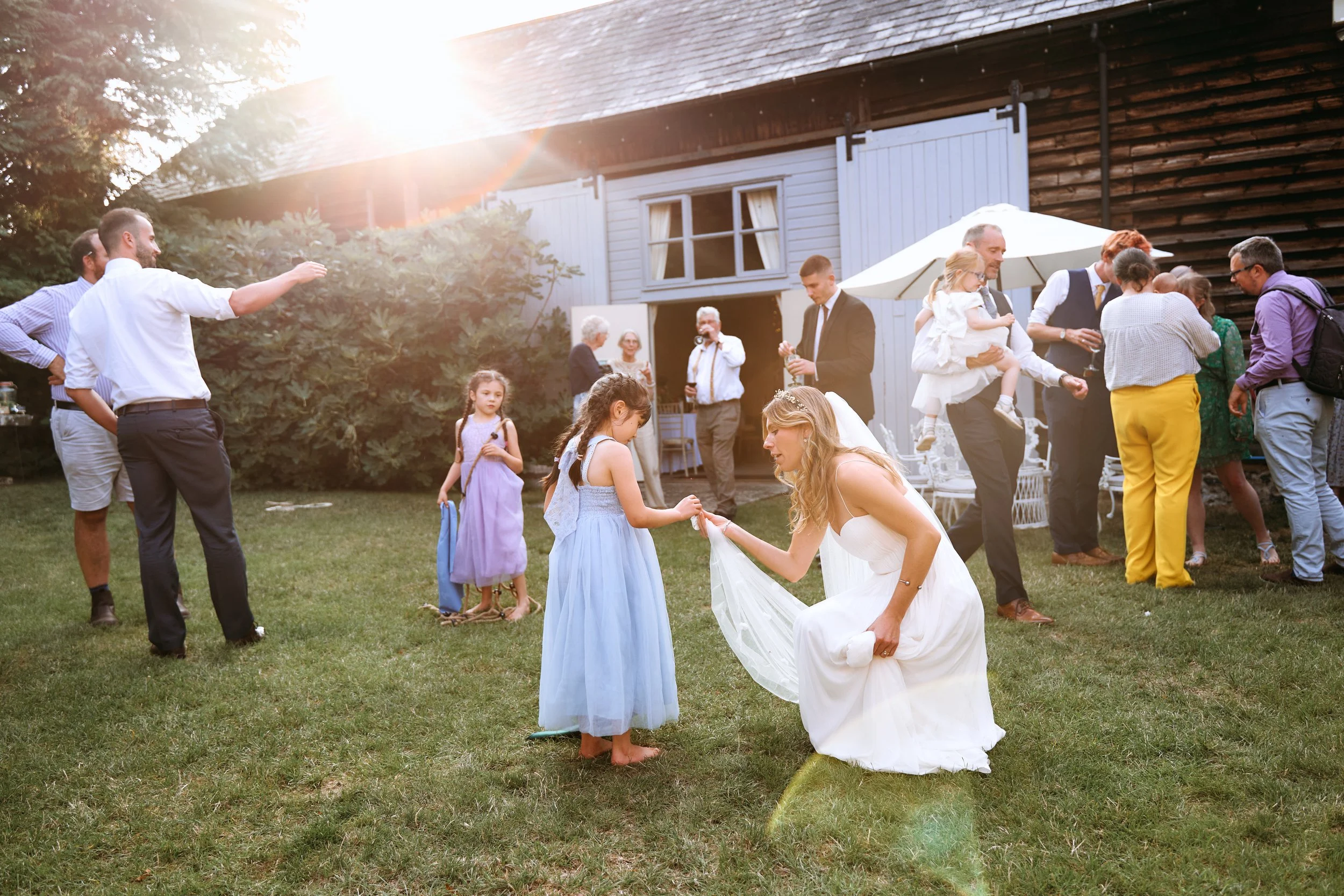 People celebrating outdoors, with a woman in a white dress and veil, likely a bride, kneeling and holding hands with a young girl in a light blue dress, during a wedding reception in the late afternoon sunlight.