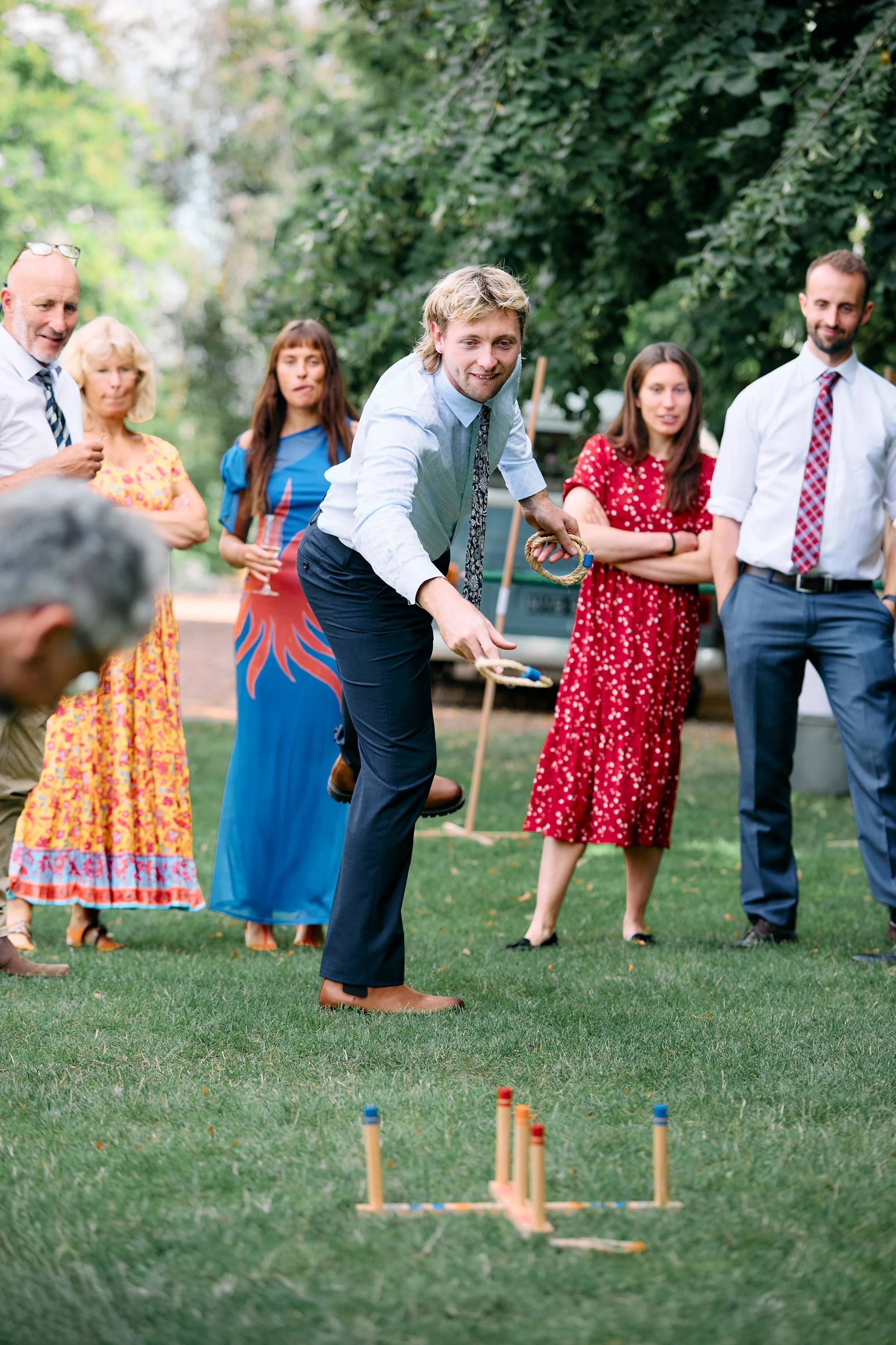 A group of people gathered outdoors on grass, watching a man in a shirt and tie playing a game with small sticks and colored markers on the ground.