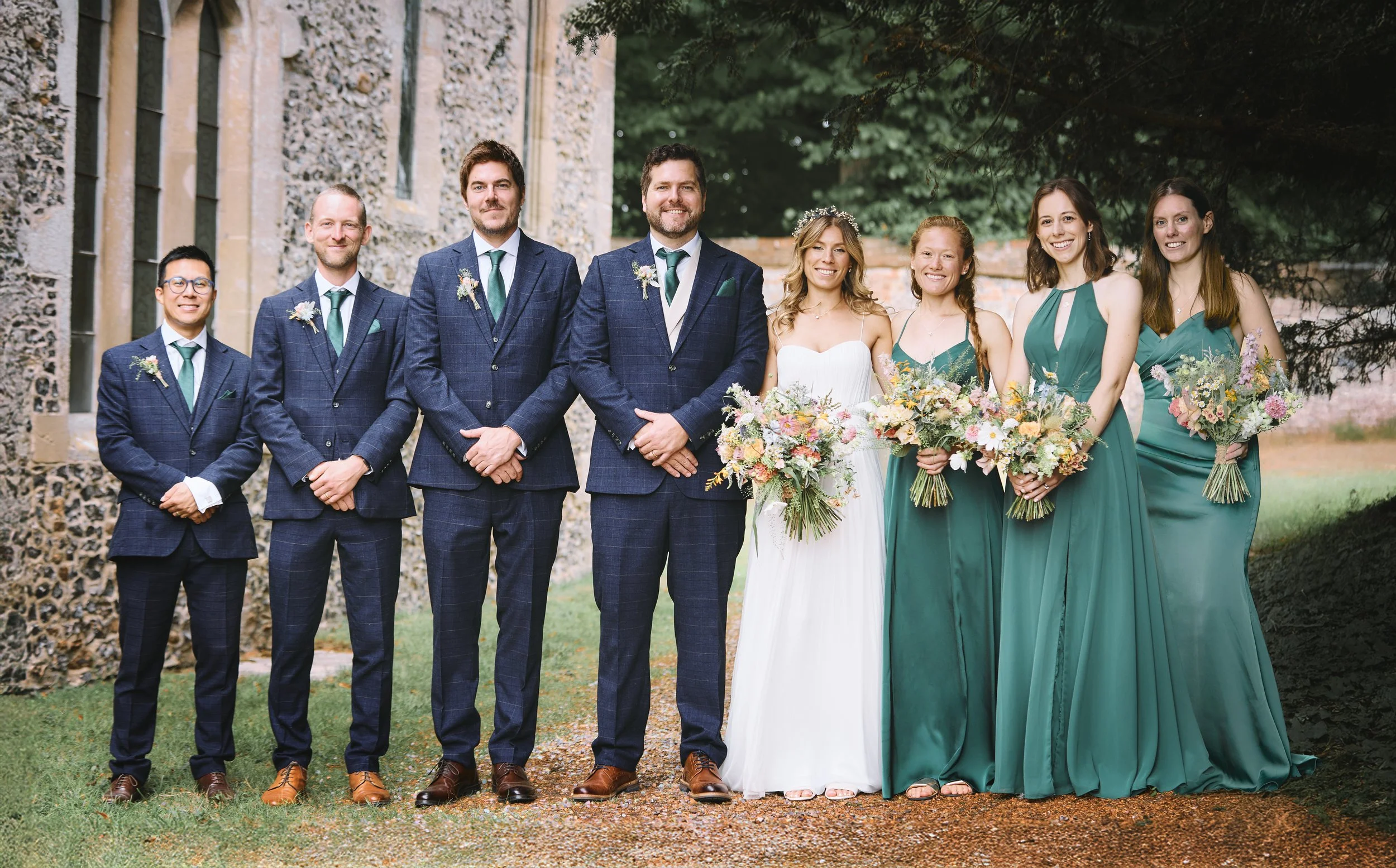 A group of nine people dressed in formal attire, standing outdoors in front of an old stone building and trees, for a wedding celebration. The bride and groom are at the center, with bridesmaids and groomsmen on either side, all smiling and holding b