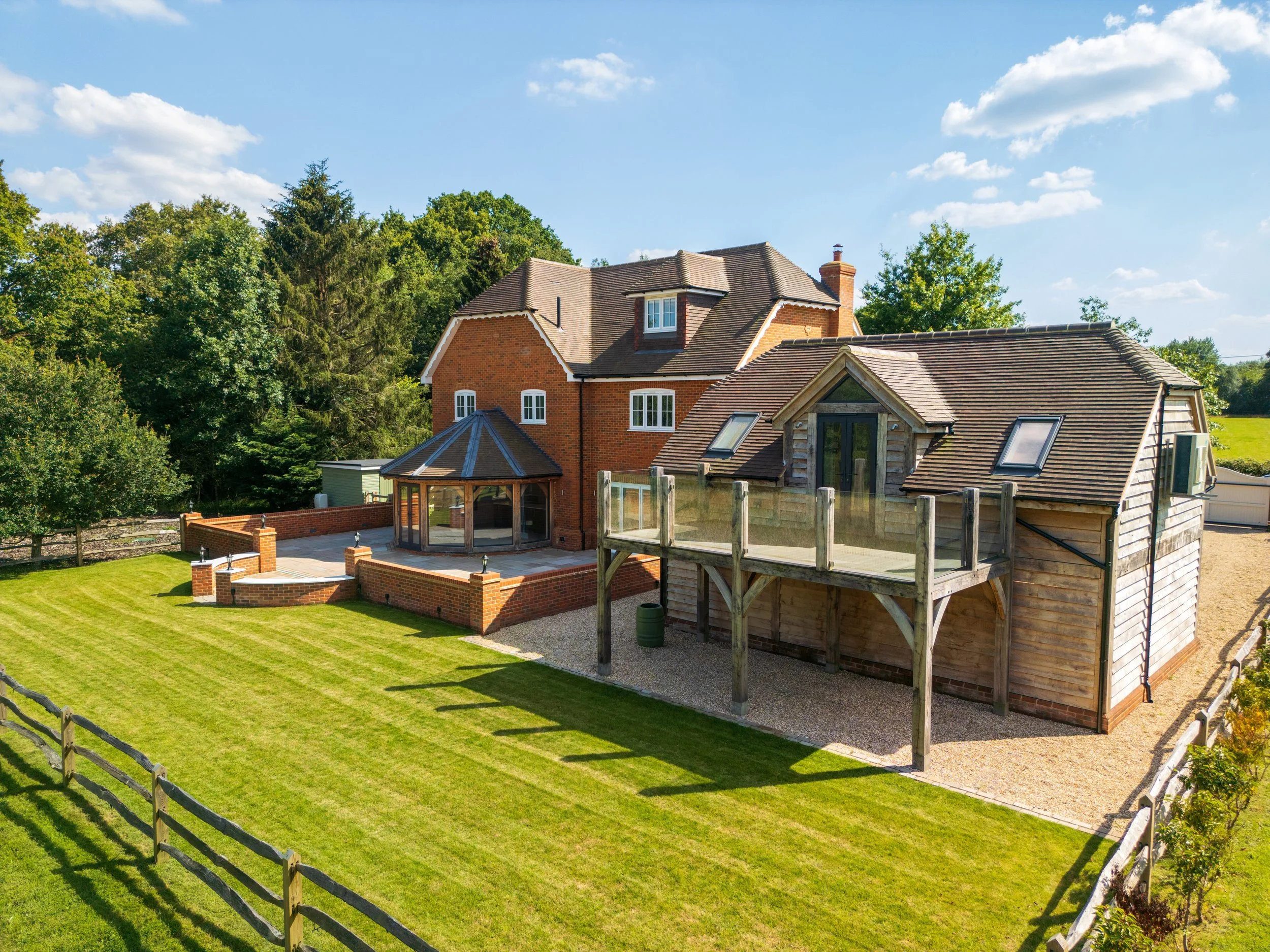 A large house with brick and wood exterior, a glass extension, a balcony, and a well-maintained grassy yard surrounded by a wooden fence, with trees and a blue sky in the background.
