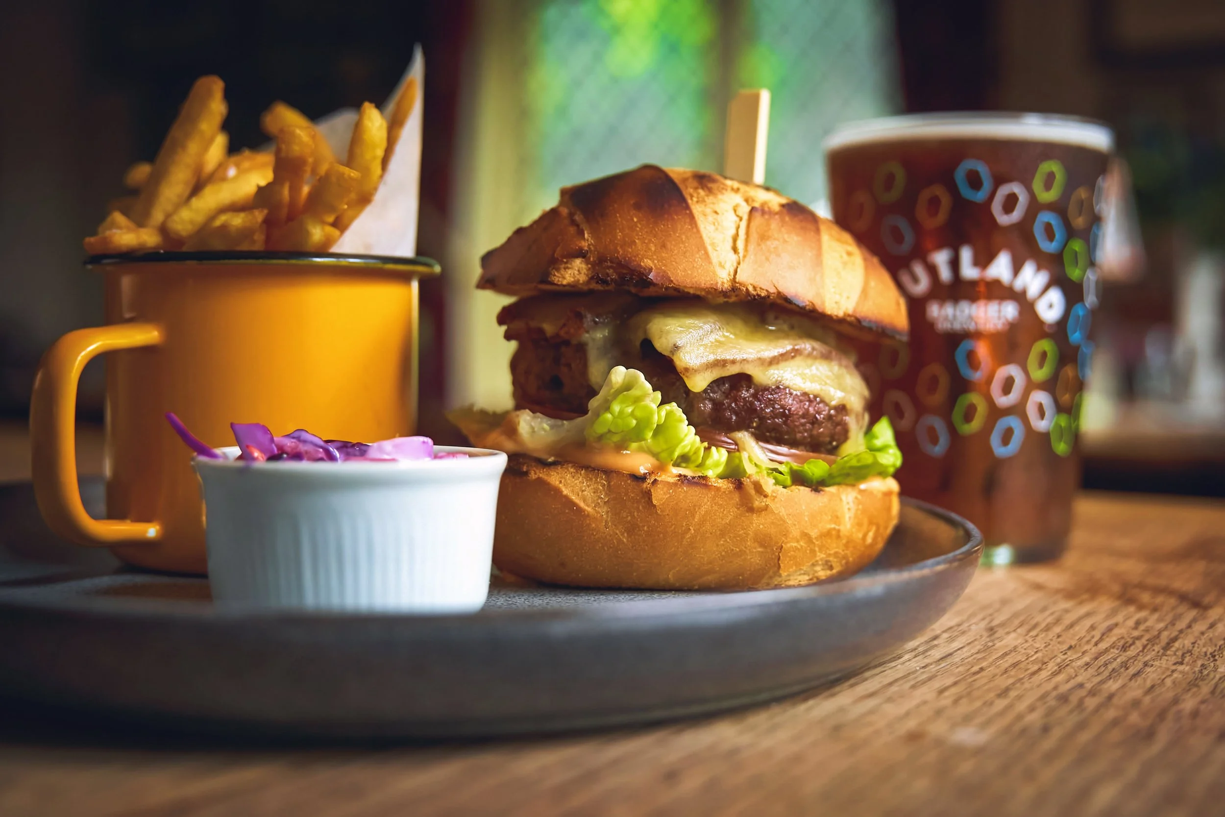 A plate with a cheeseburger, French fries in an orange cup, a small cup of pink and purple coleslaw, and a cup of soda from Outland Brewery on a wooden table.