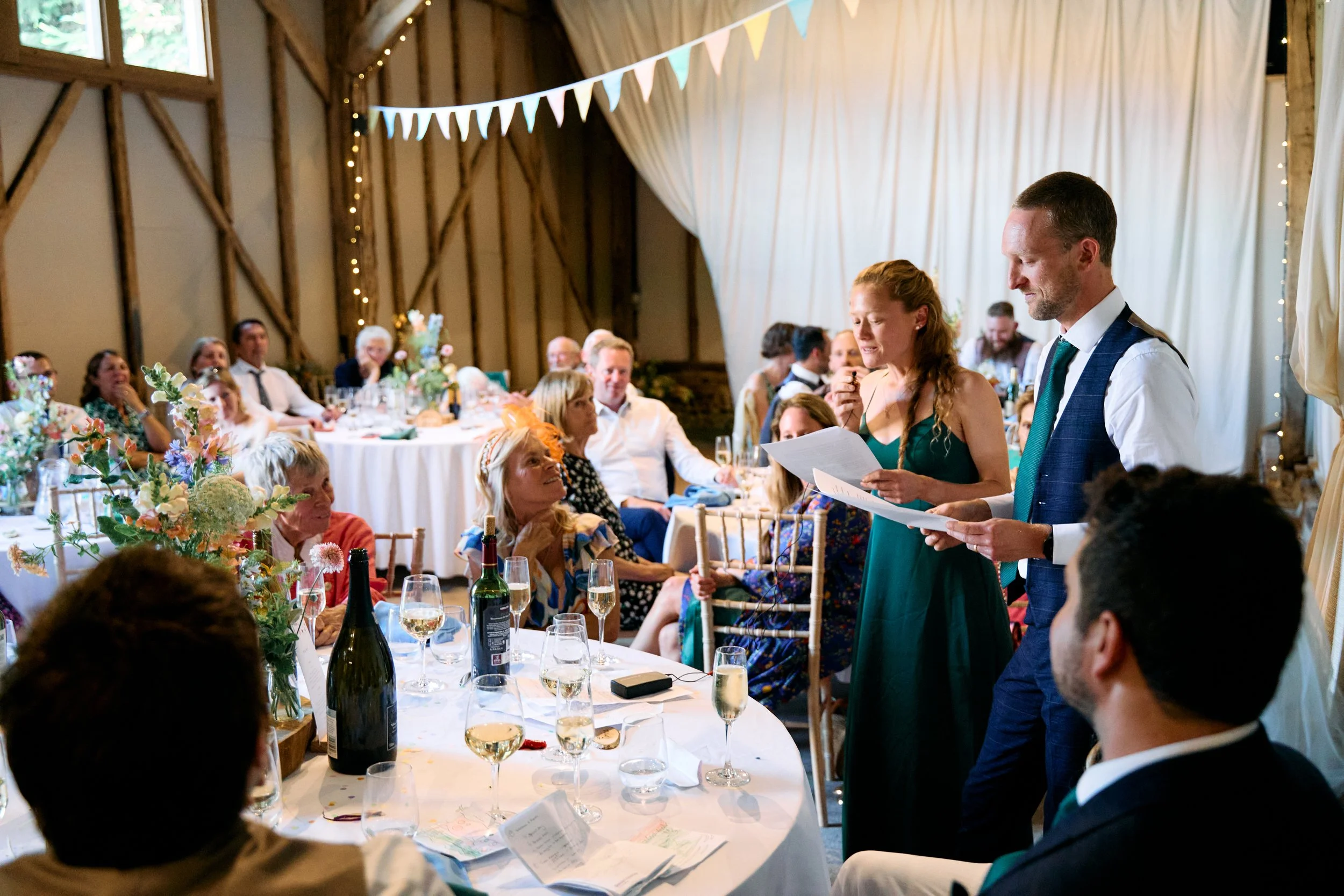 Wedding reception inside a barn with a couple giving a speech, surrounded by seated guests at decorated tables with flowers, wine bottles, and glasses, white drapery and bunting hanging from the ceiling.