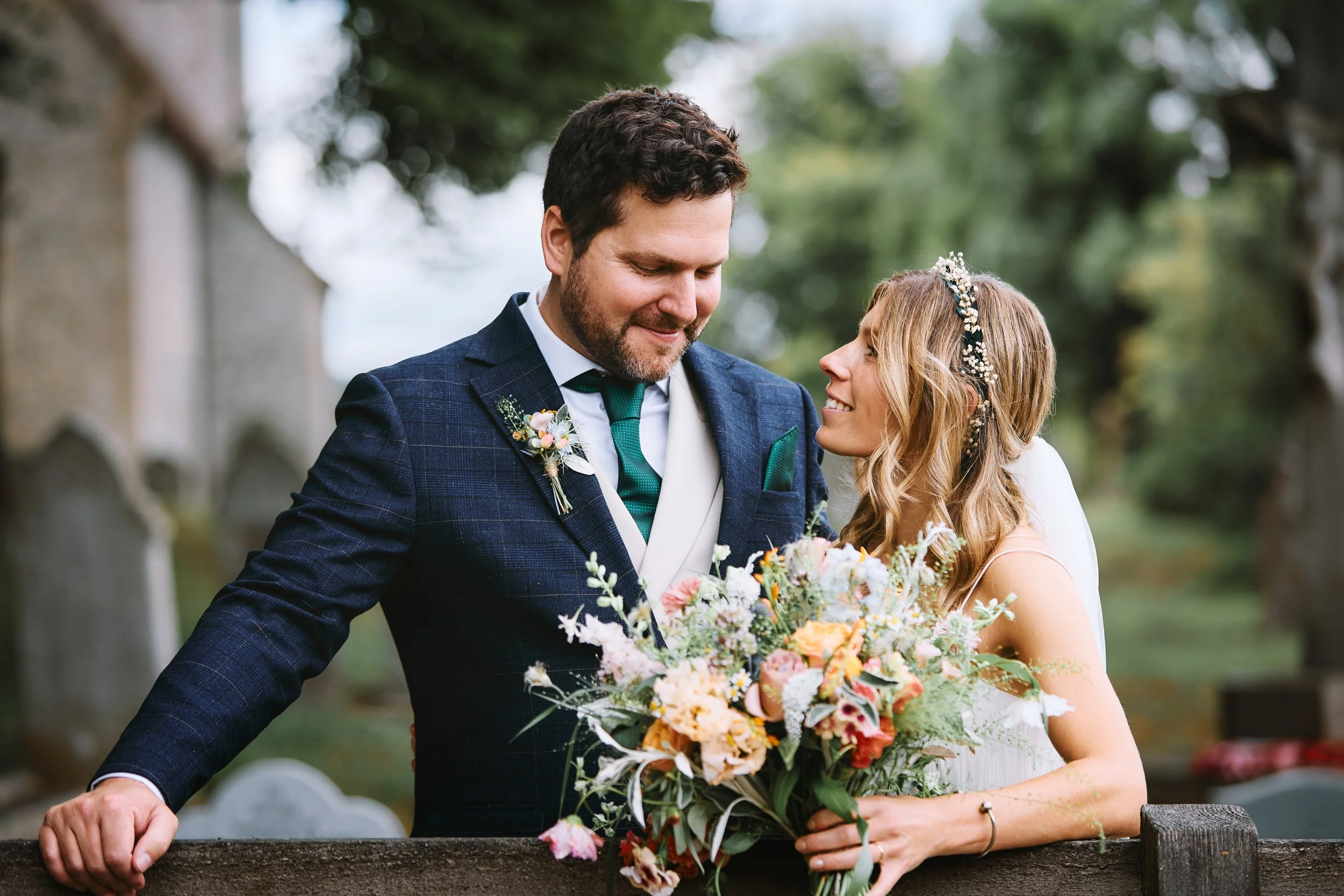 A bride and groom stand close together, smiling at each other, outdoors near a stone fence, with greenery and trees in the background. The bride holds a colorful bouquet and wears a floral headband, while the groom is dressed in a navy suit with a bo