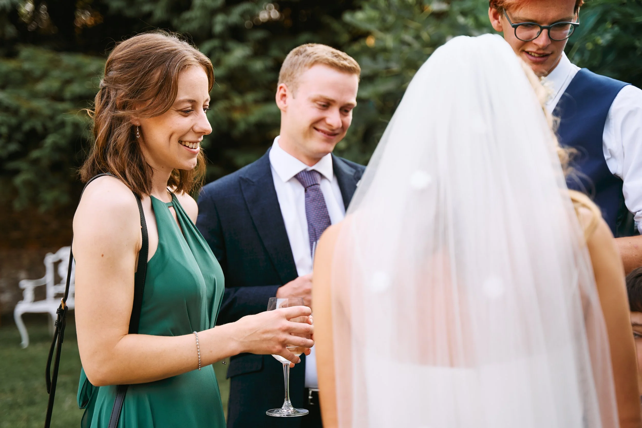 A group of people at a wedding reception, including a woman in a green dress holding a glass of champagne, a man in a suit and tie, and others, with a bride in a white veil.
