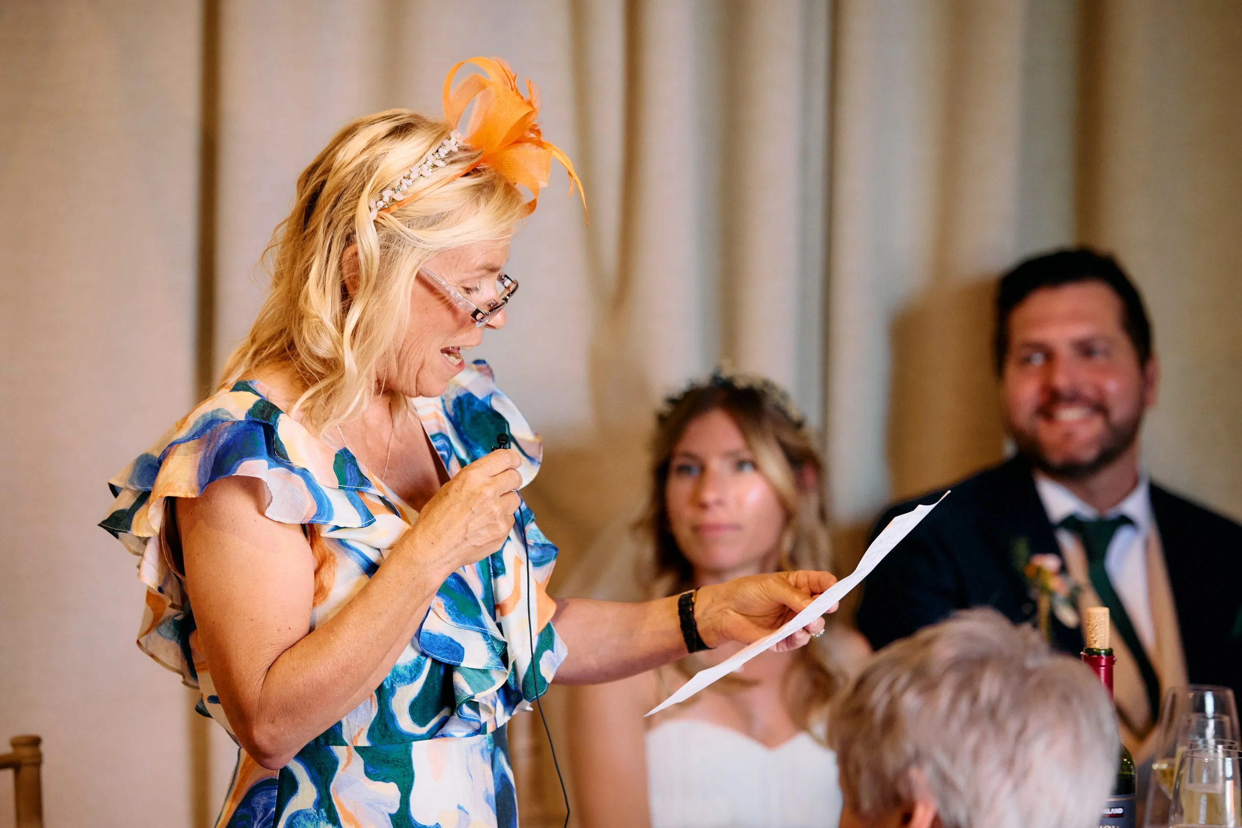 A woman dressed in a colorful dress and wearing orange fascinator hat reads her speech at a wedding reception, with a bride and groom sitting nearby, and other guests visible in the background.