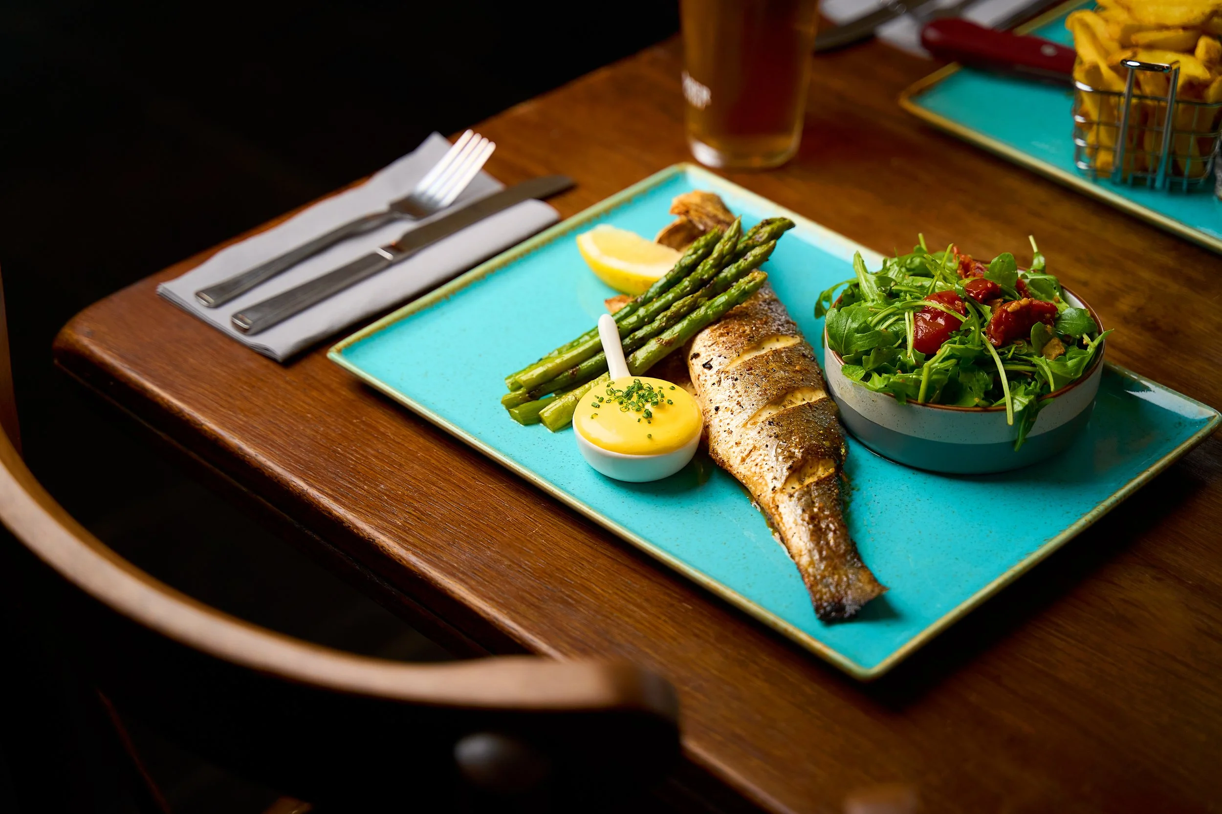 Grilled fish with asparagus, lemon wedge, and a bowl of salad on a turquoise ceramic plate with a small cup of sauce and a fork and knife on a white napkin on a wooden table.