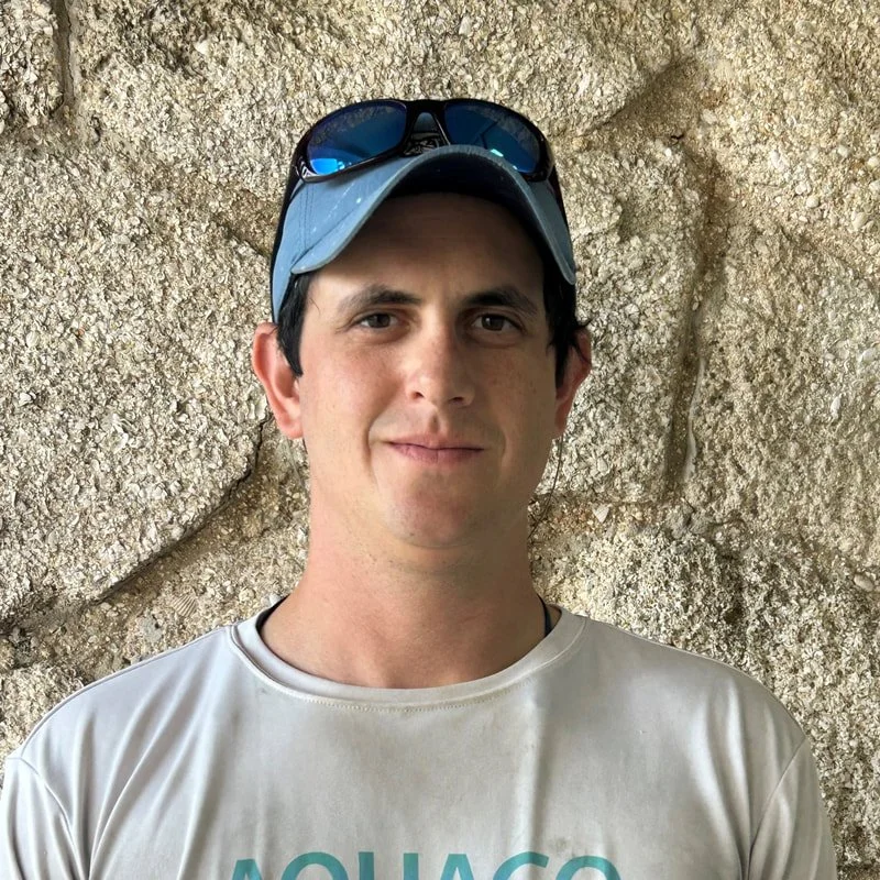 A young man wearing a gray baseball cap with sunglasses on top, standing in front of a rocky background.