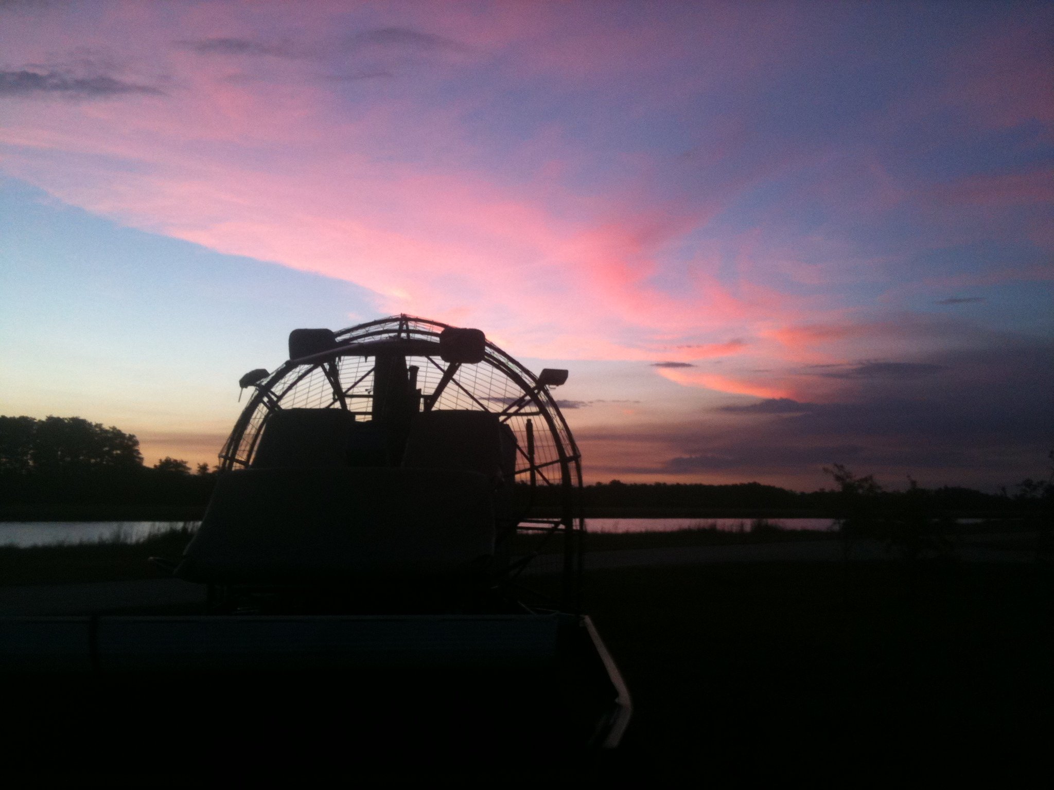 Silhouetted airboat against a colorful sunset with pink and purple clouds over a body of water and distant trees.