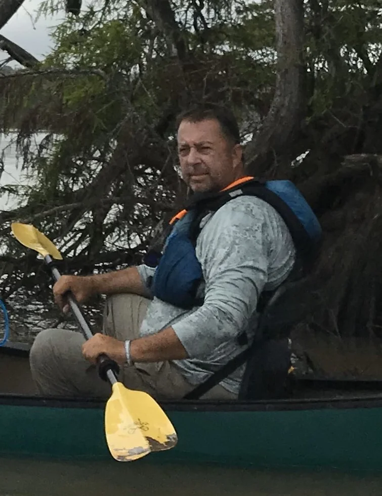 A man in a kayak holding a yellow paddle, wearing a gray long-sleeve shirt and a blue life vest, sitting on the water near a tree with dense branches.