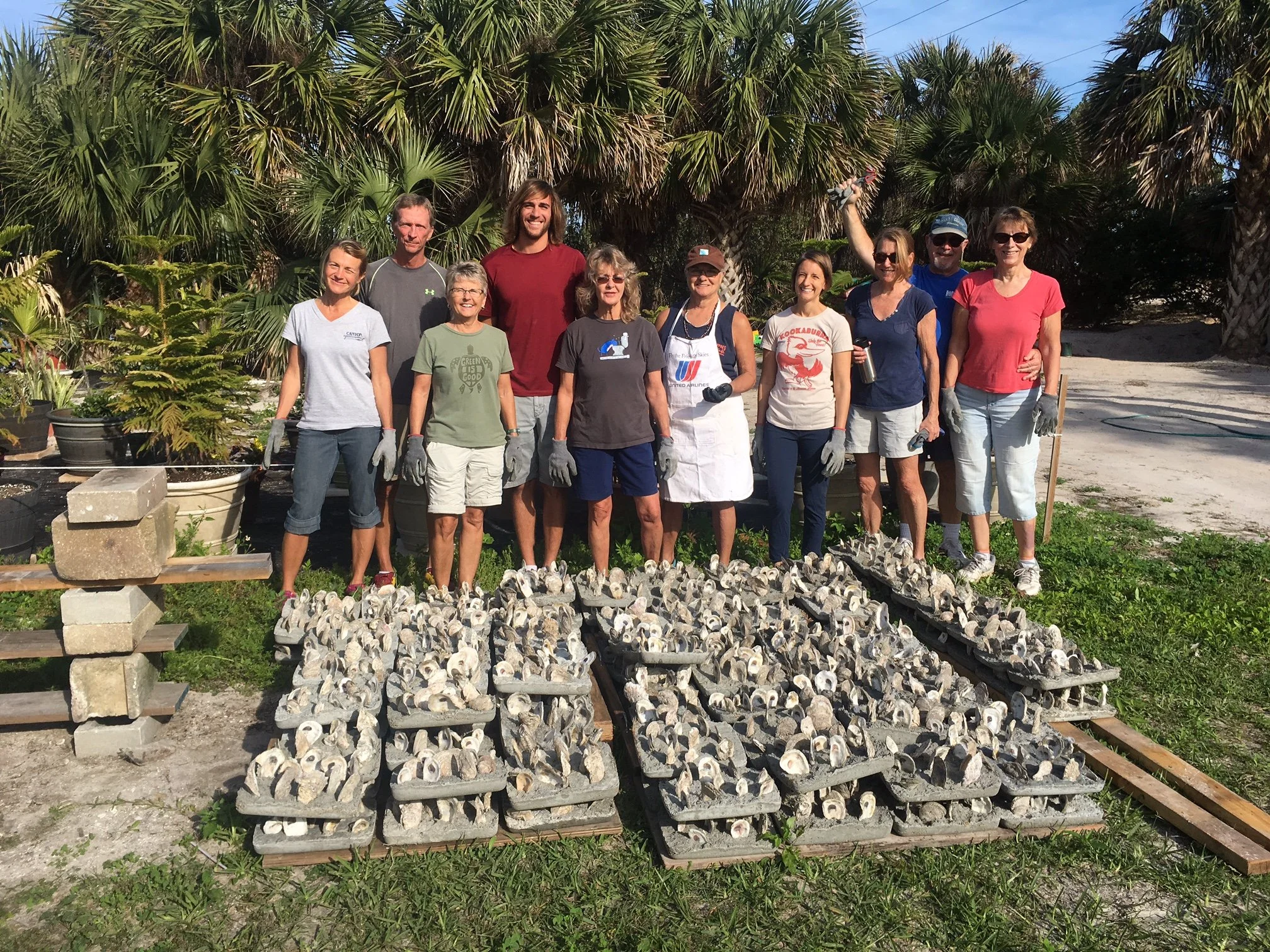 Group of people standing outdoors in front of palm trees, wearing gardening gloves, with trays of seashells or similar items laid out on the ground in front of them.