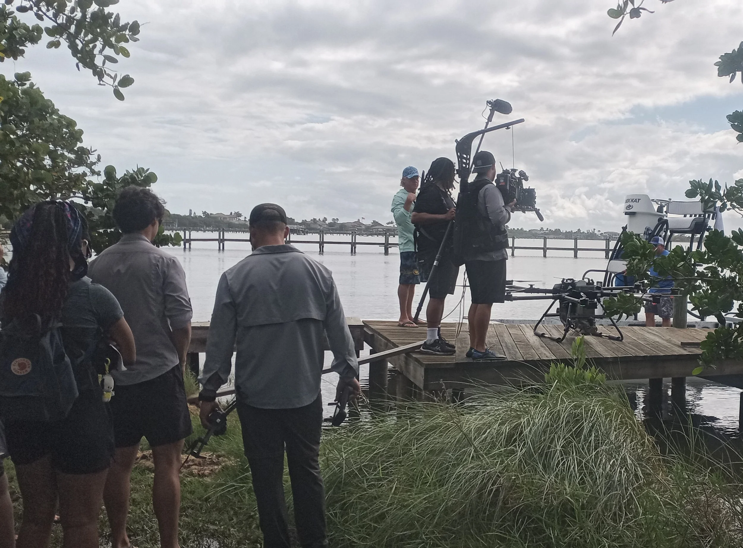 Filmmakers and crew filming by a river with a bridge and cloudy sky in the background.