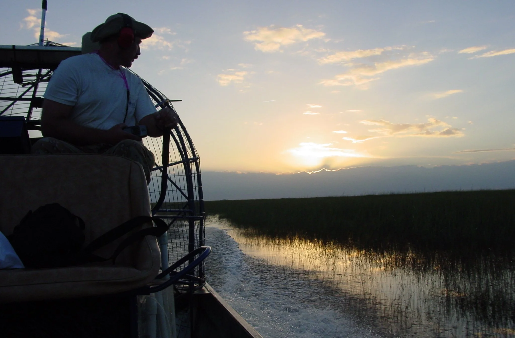 A person sitting on a boat at sunset, wearing a hat and headphones, navigating through a waterway with tall grasses on either side.