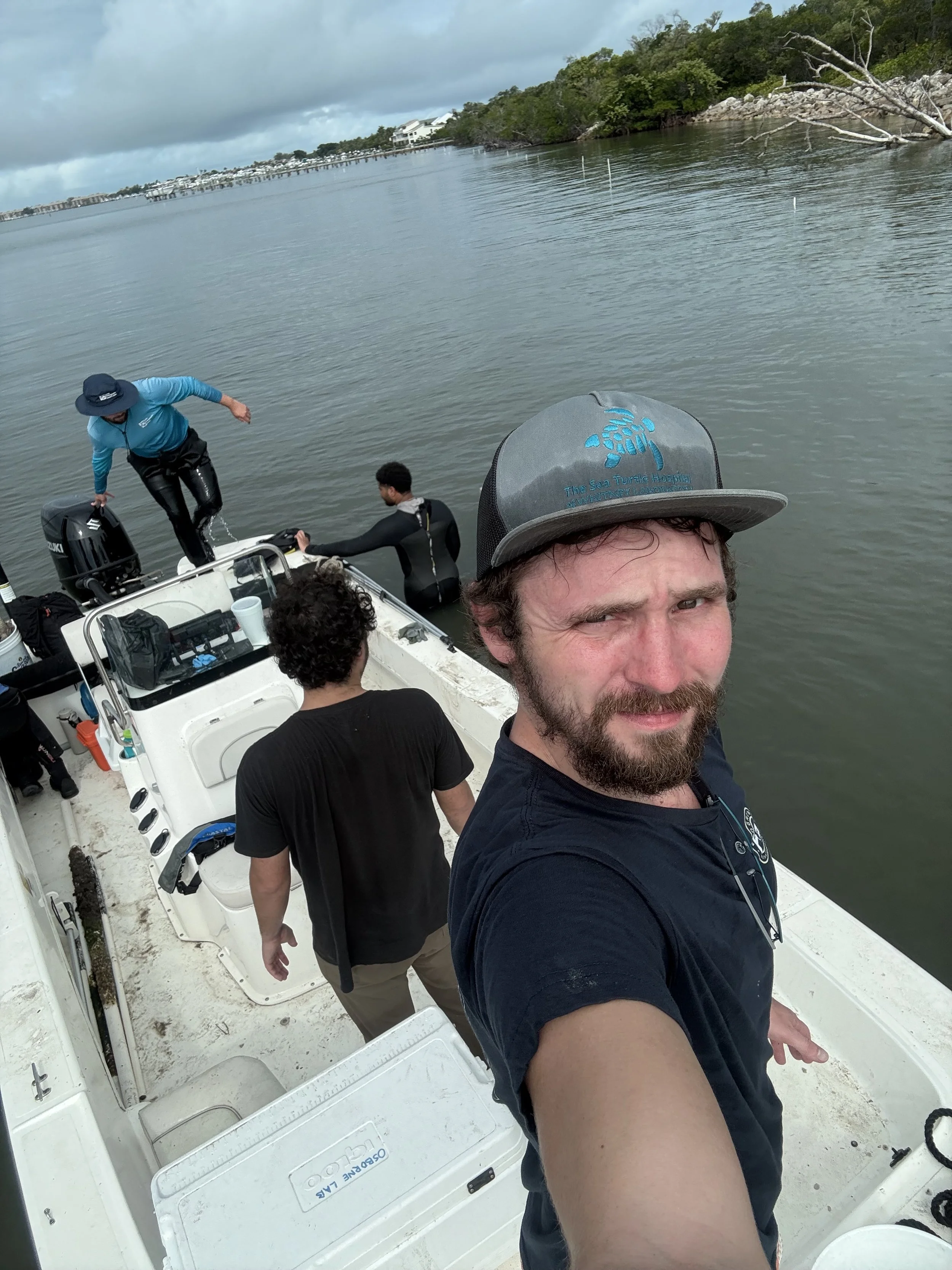 A man with a beard taking a selfie on a boat with other people, some are sitting and others are boarding the boat, on a body of water near a green shoreline.