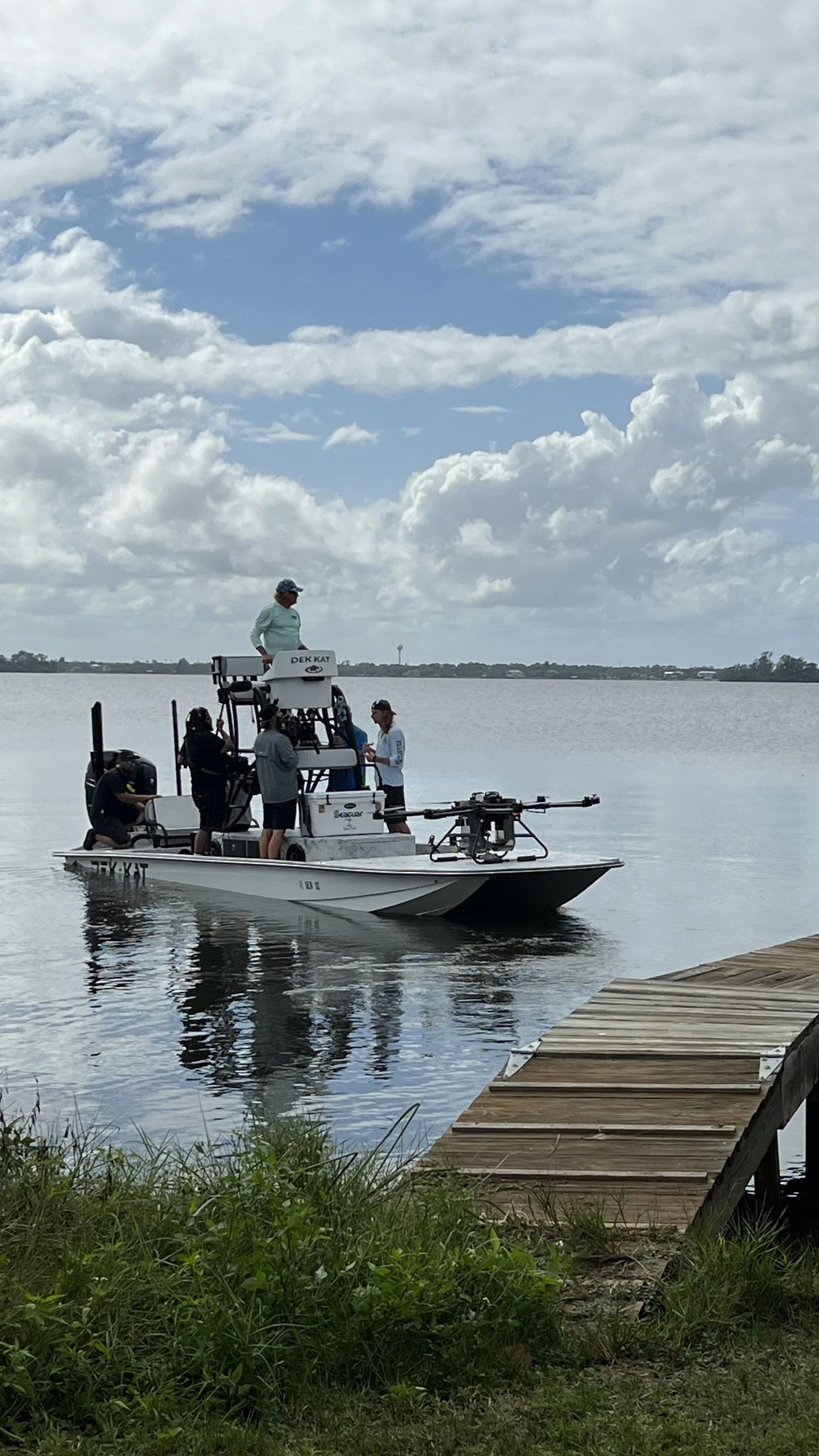 A small boat on a lake with four people onboard operating a drone, docked near a wooden pier, with a partly cloudy sky overhead.