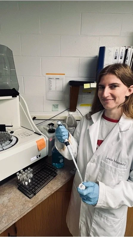 A woman in a white lab coat and gloves holds a pipette in a laboratory setting with scientific equipment and test tubes on the counter.