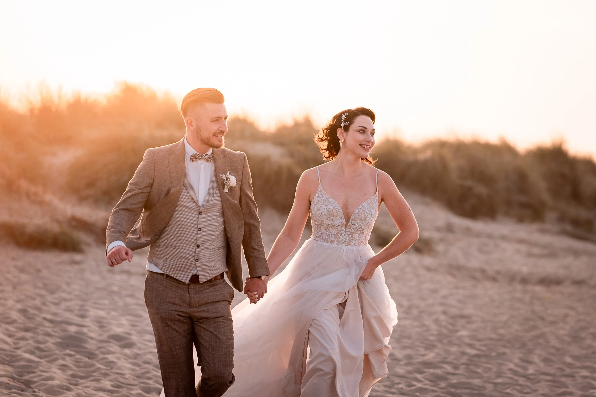 Ein Hochzeitspaar läuft Hand in Hand am Strand bei Sonnenuntergang, die Braut trägt ein fließendes, elegantes Brautkleid mit Spitze und die Haare sind dekoriert, der Bräutigam trägt einen braunen Anzug mit Krawatte.