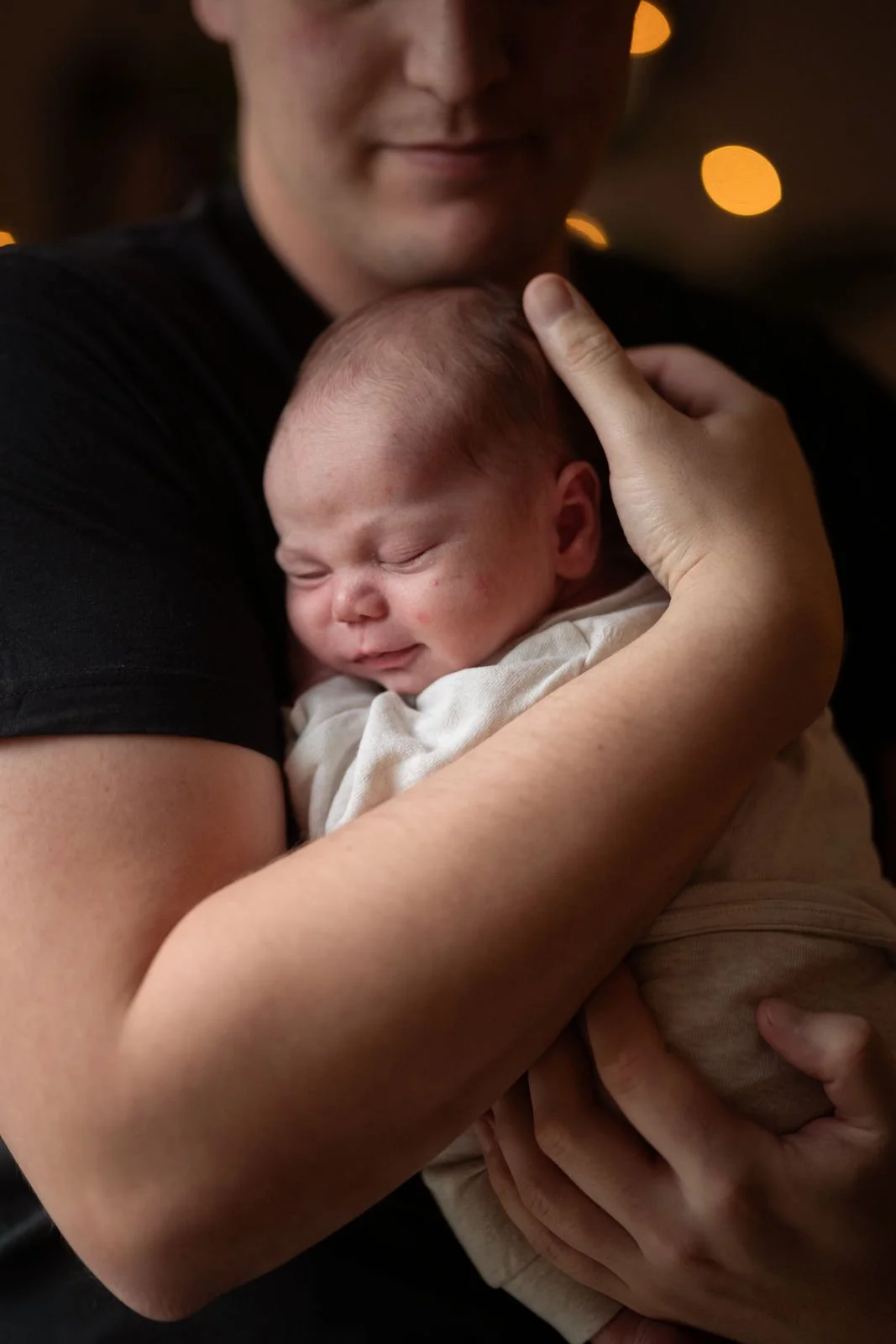 Ein Mann hält ein schlafendes Baby eng an sich, die Szenerie ist warm und ein paar unscharfe Lichter sind im Hintergrund sichtbar.