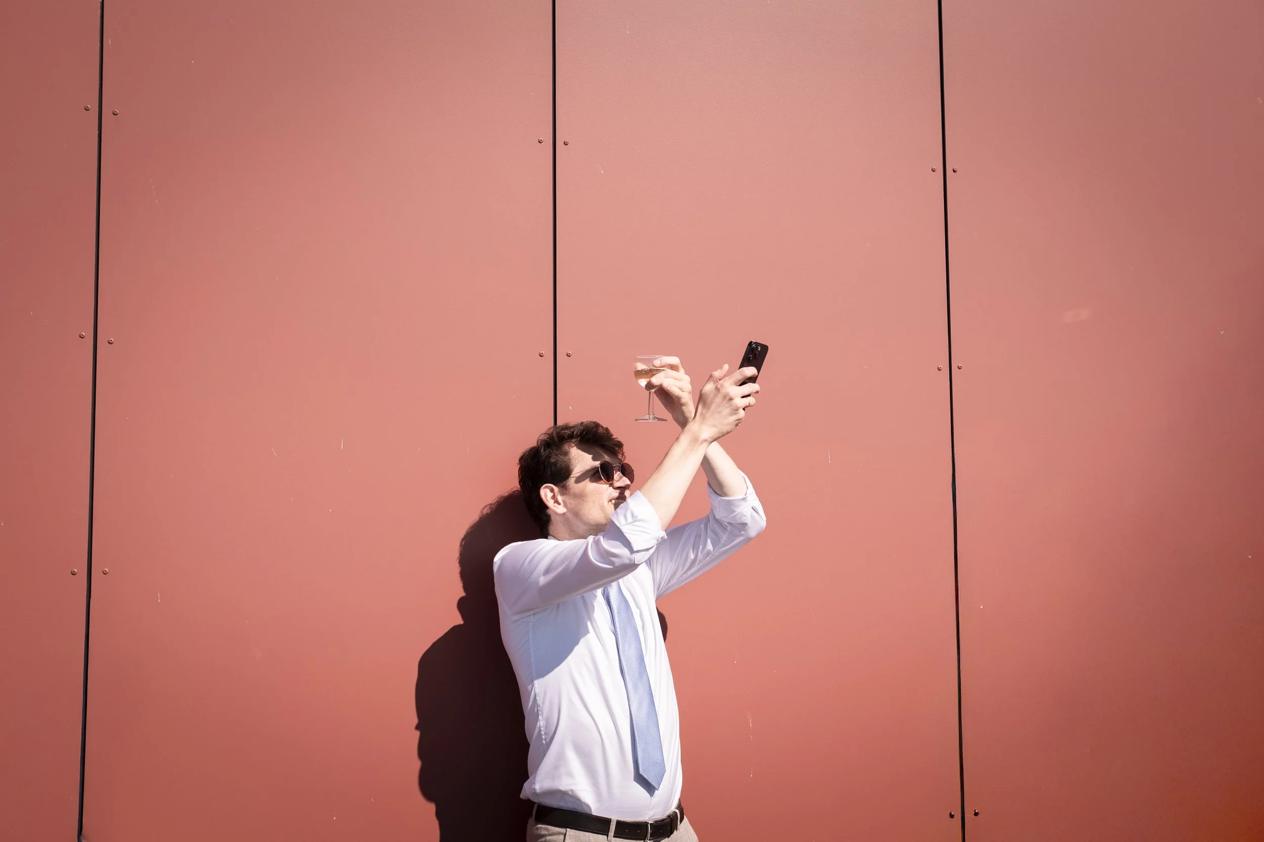 Ein Mann steht vor einer rosa Wand, trägt Sonnenbrille, weiße Hemd und Krawatte, hält ein Smartphone und ein Weinglas und macht ein Selfie.
